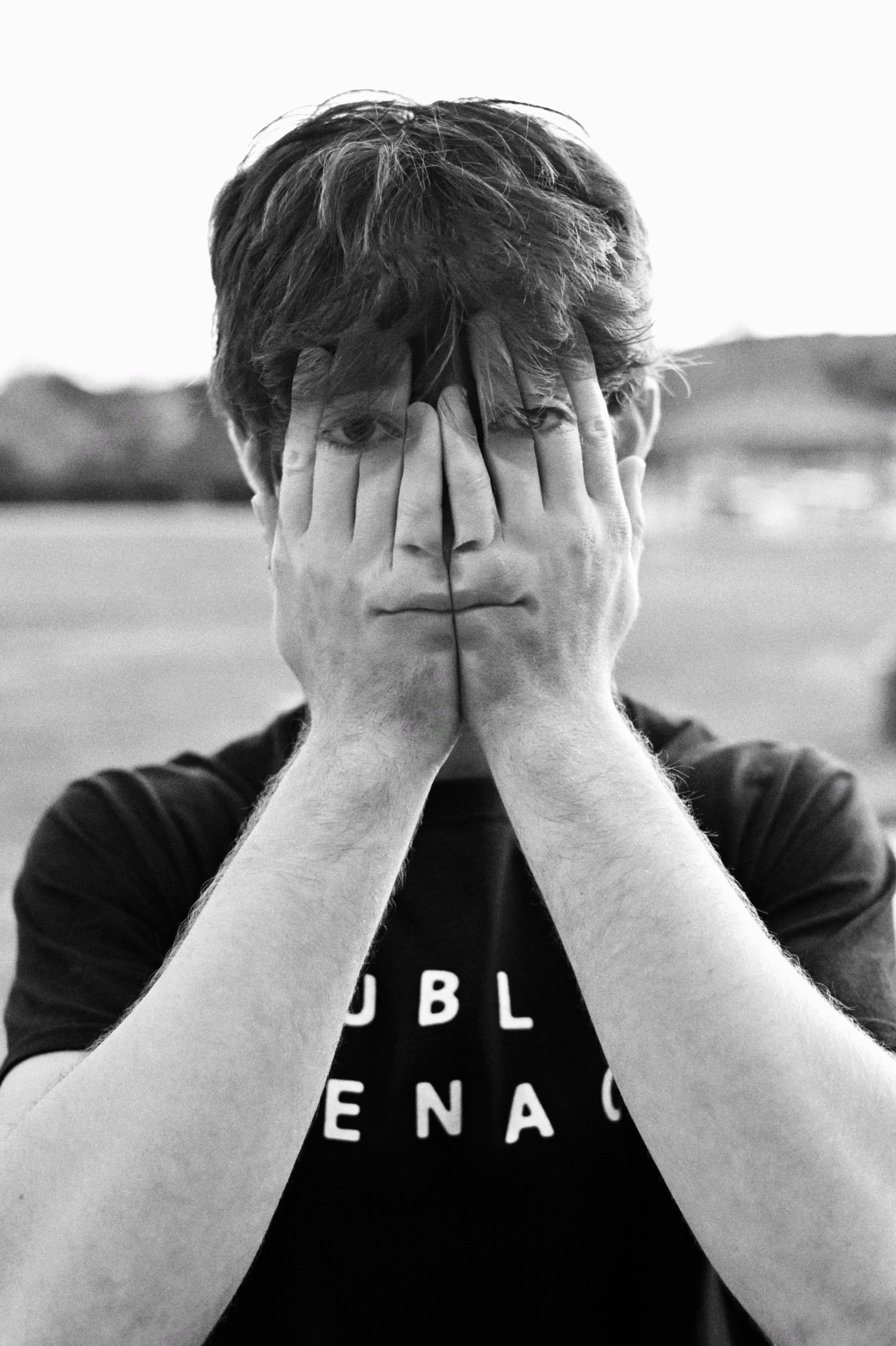 A young man with light skin and messy hair covering his eyes with his hands, outdoors with a blurred background.