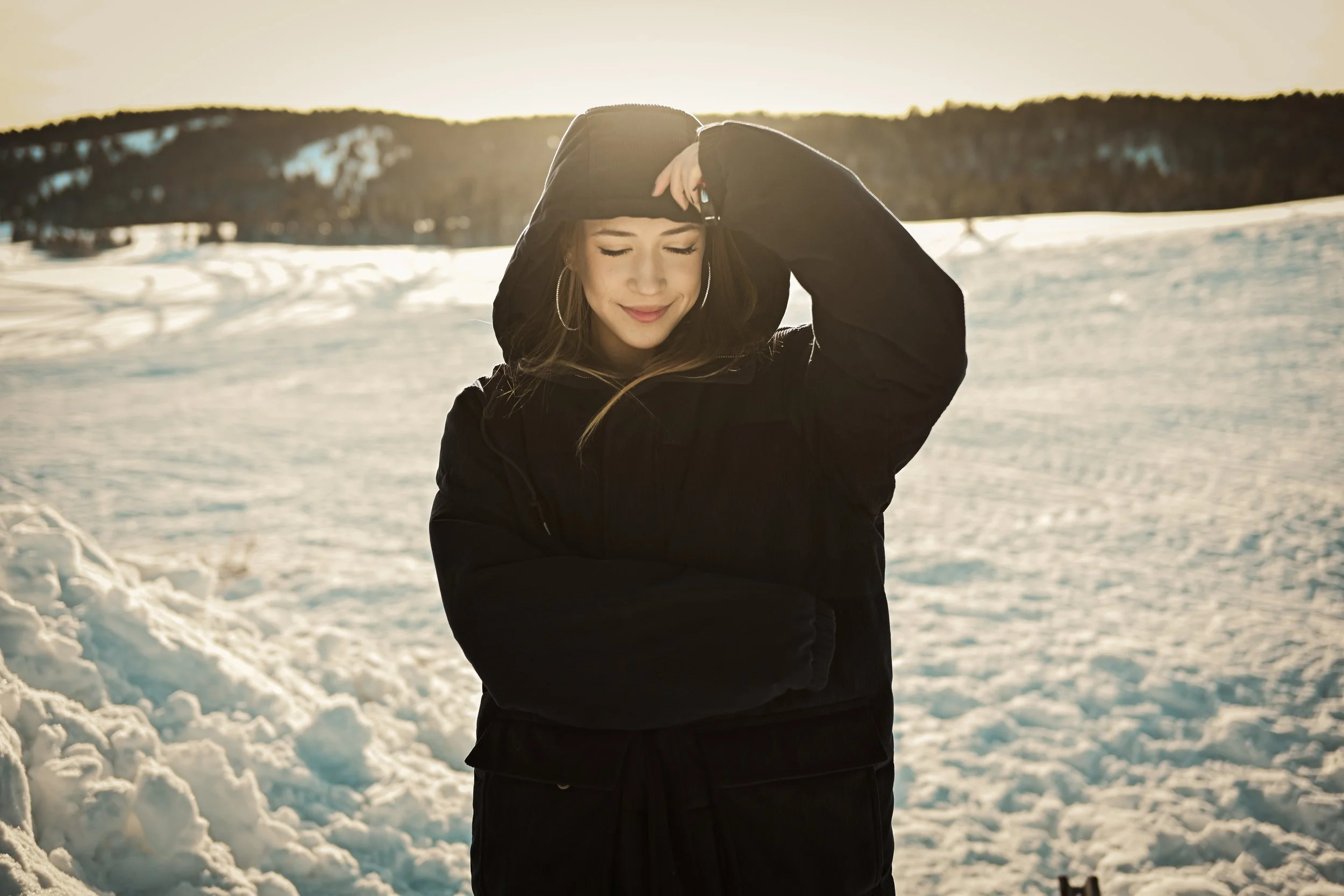 Young woman in black winter coat with hood in snowy landscape, smiling with eyes closed, during sunset or sunrise.