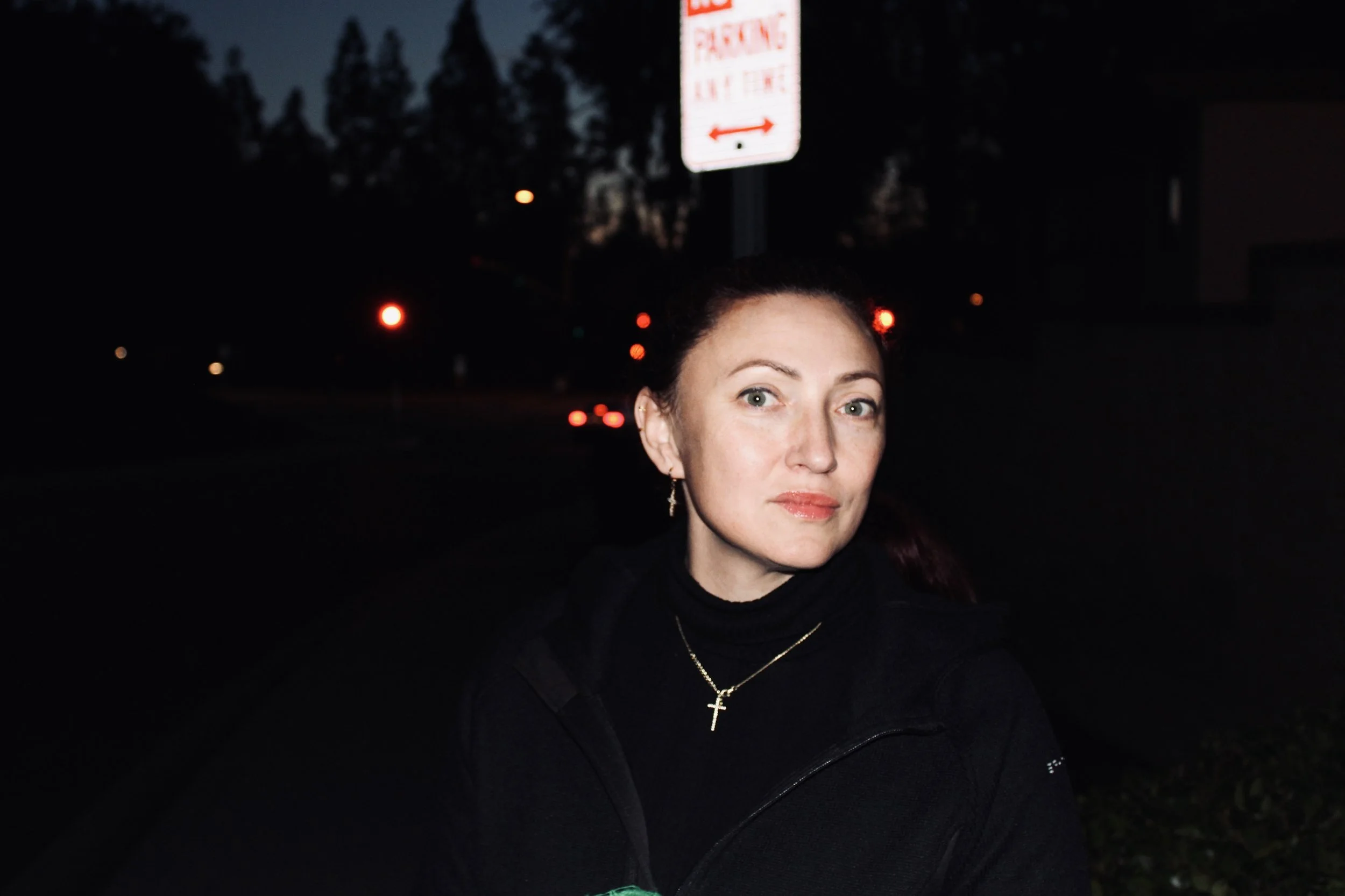 A woman with red hair and light skin wearing a black jacket, gold cross necklace, and earrings, stands outdoors at night near a street with red traffic lights in the background. There is a traffic sign above her. The sky is dark, and trees are faintly visible.