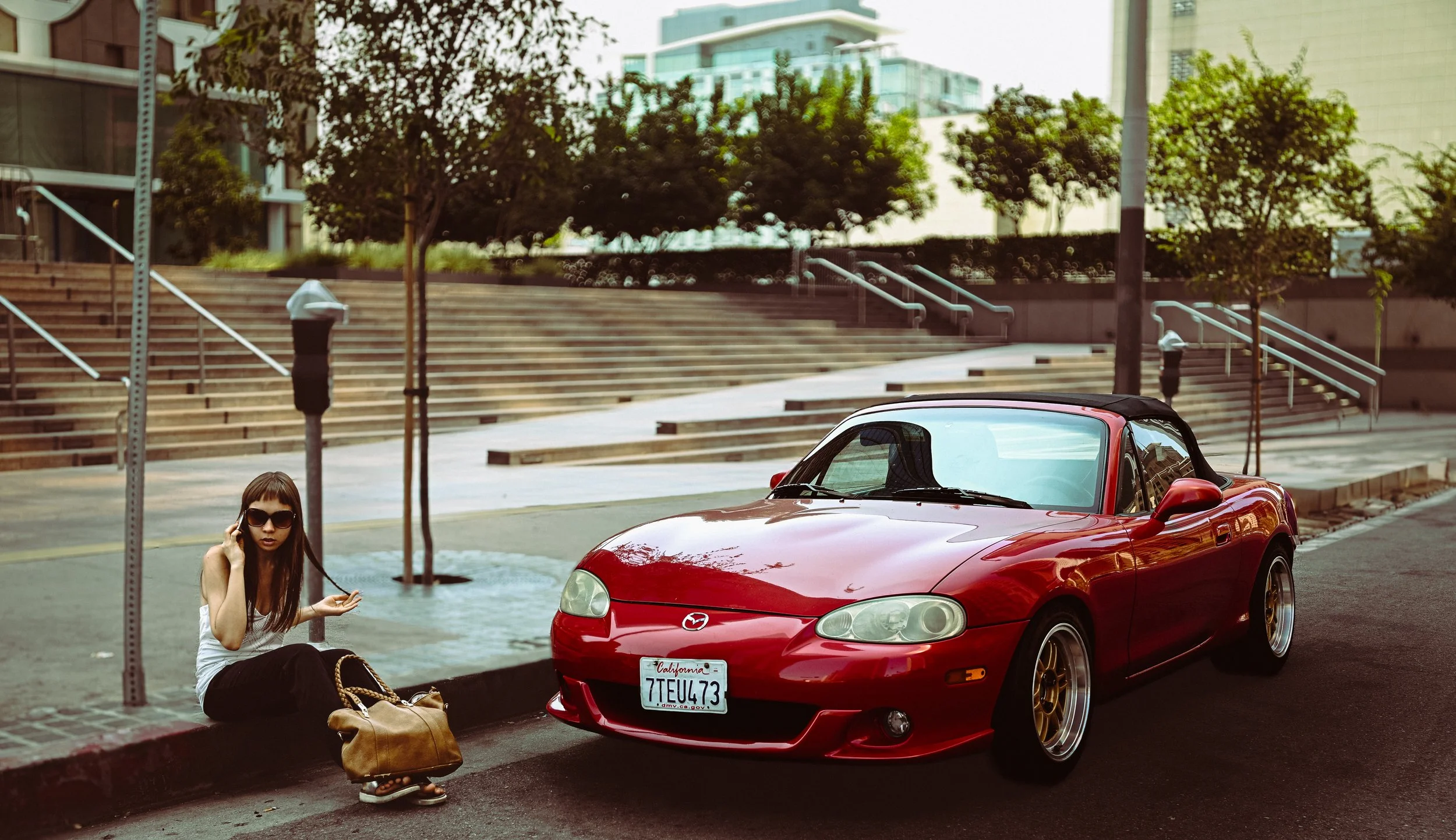 A woman sitting on the sidewalk next to a red convertible Mazda MX-5 Miata in an urban area with stairs, trees, and modern buildings in the background.