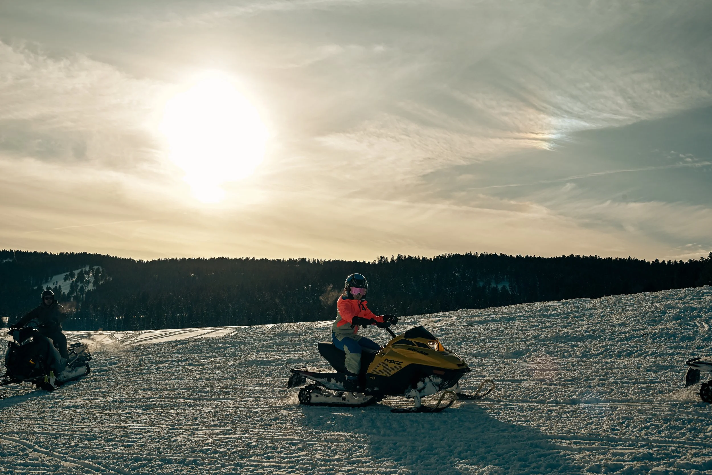 Person riding a yellow snowmobile on snow-covered terrain with another snowmobile and rider in the background, under a partly cloudy sky with the sun shining.