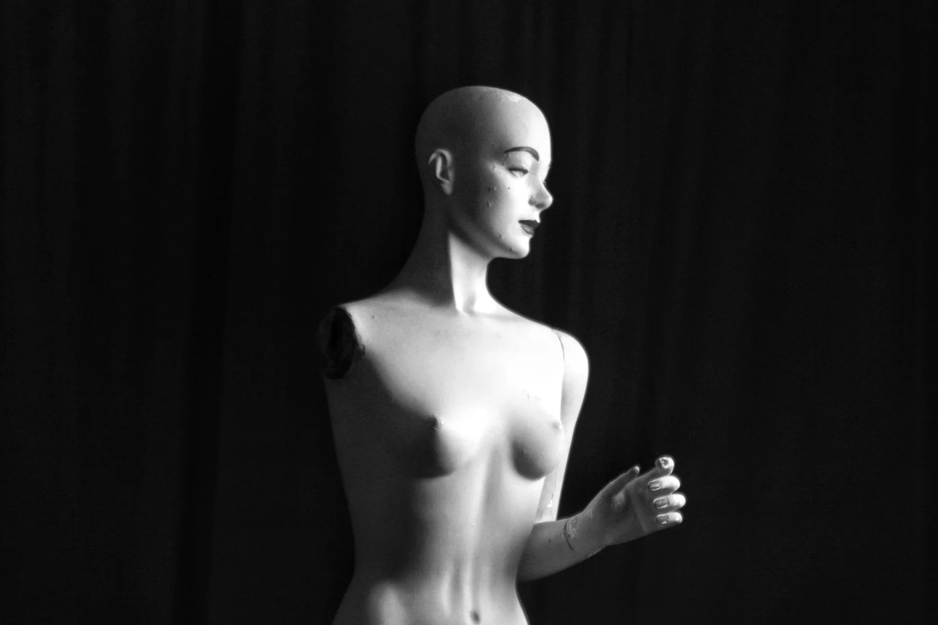 A black and white photograph of a mannequin with a smooth, featureless head and an elegant pose, set against a dark background.