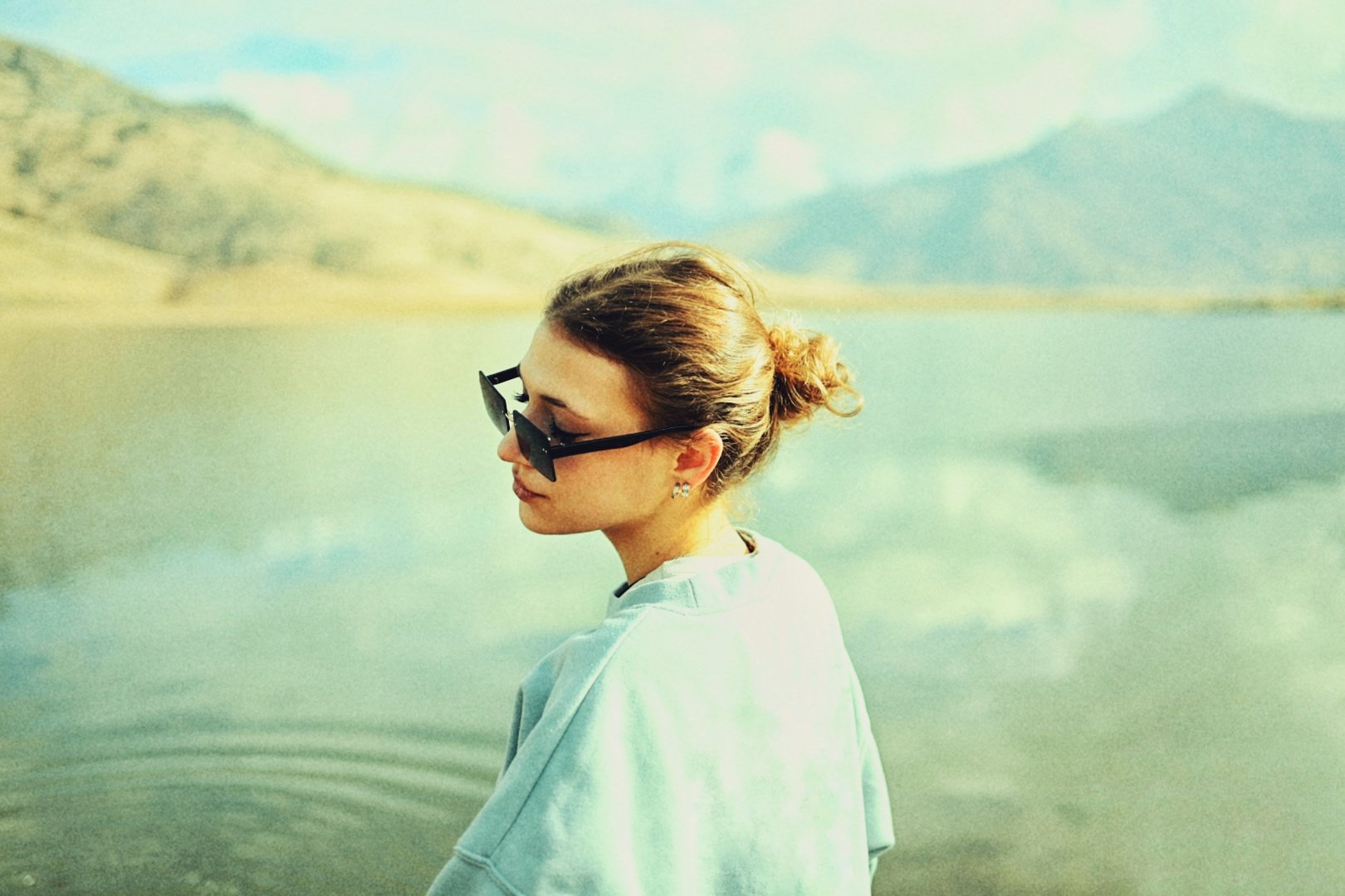 A woman with brown hair tied in a bun, wearing black sunglasses and a light-colored sweatshirt, standing near a calm lake with mountains in the background.