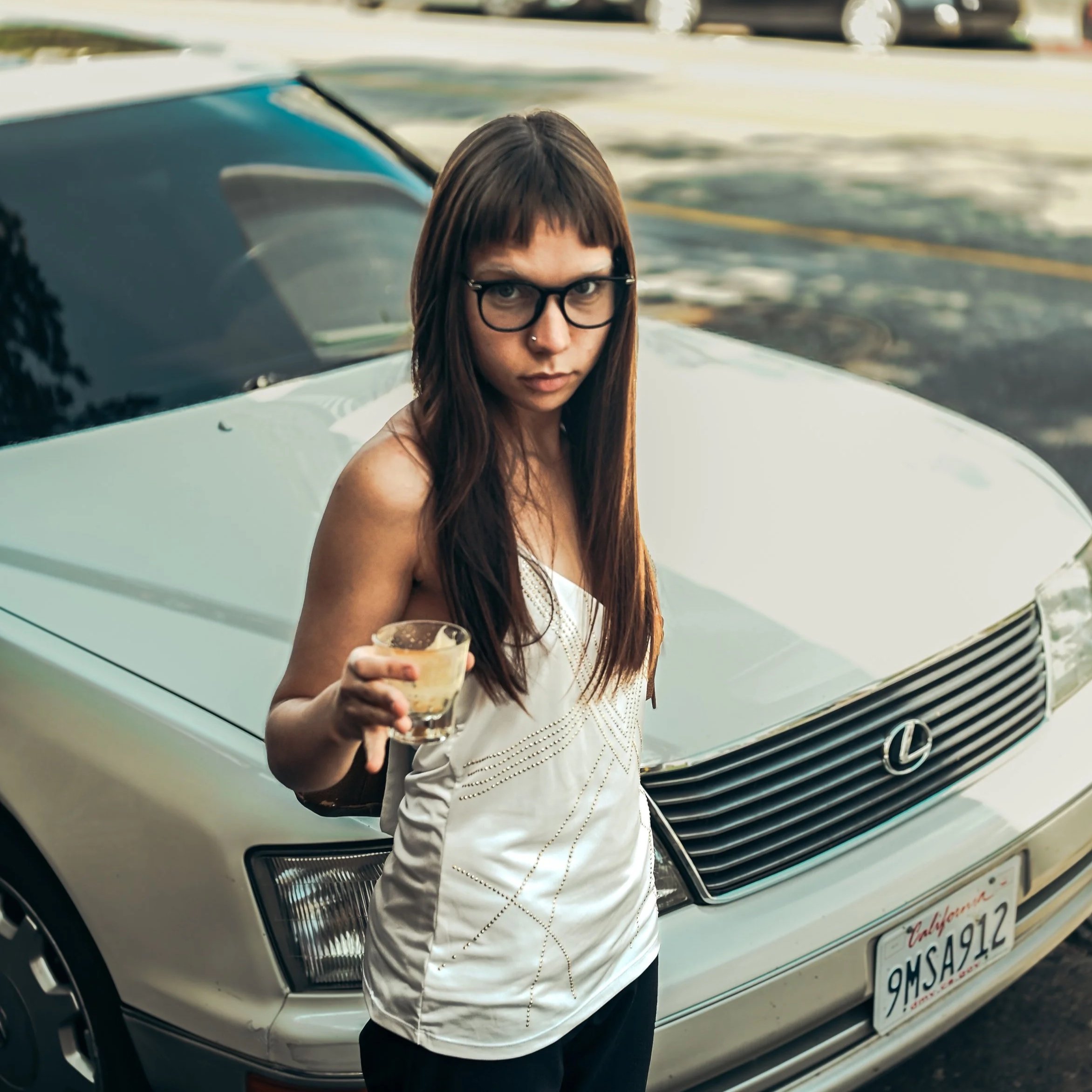 A young woman with long brown hair, black glasses, and a nose piercing holding a drink, standing in front of a white Lexus car with California license plate 9MSA912.