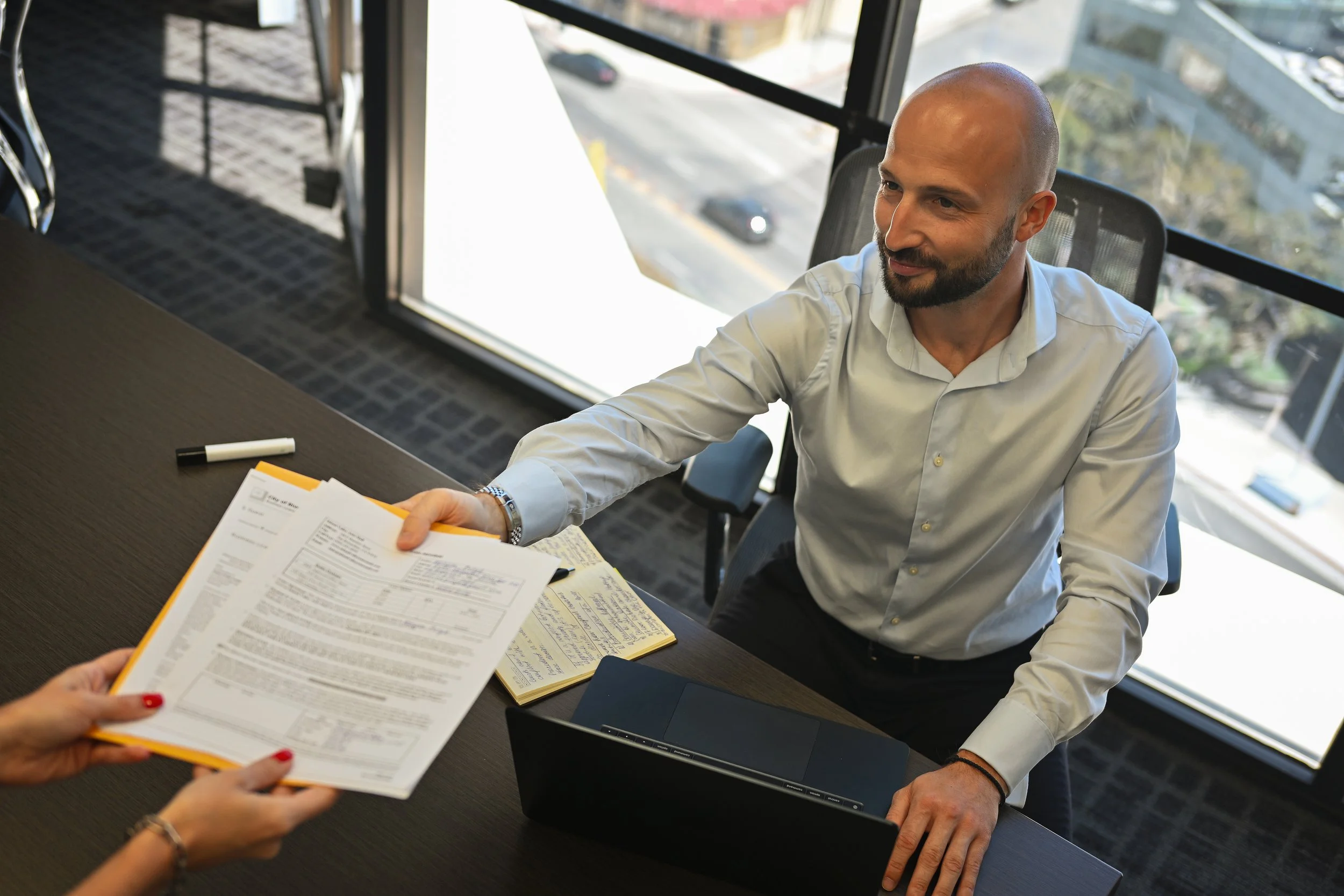 A man in a white shirt sitting at a desk, receiving a document from a person with red painted nails, in an office with large windows and a city view.