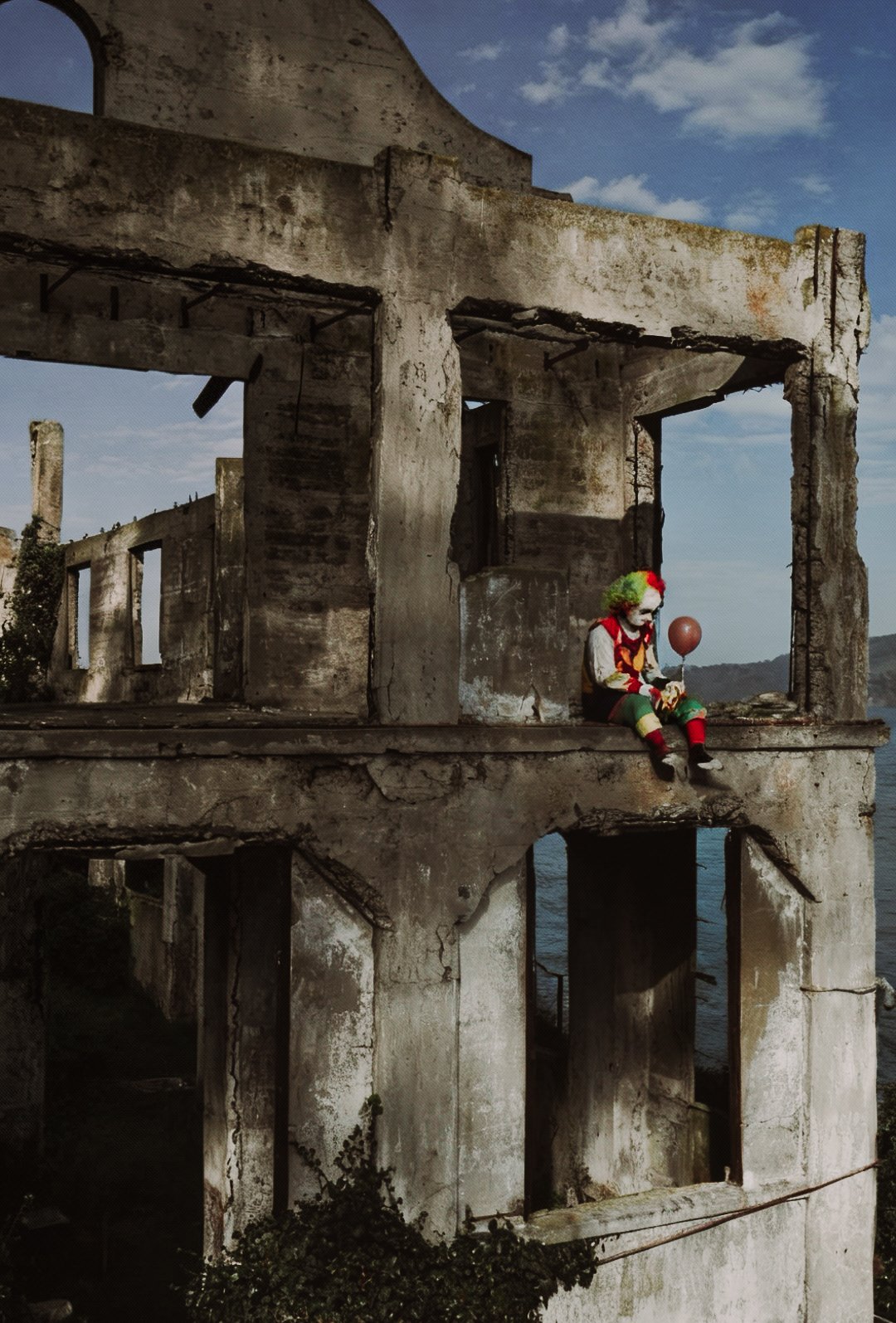 A person dressed as a clown with multicolored hair, makeup, and costume, sitting on a ledge of a dilapidated, ruined building, holding a balloon and looking down.