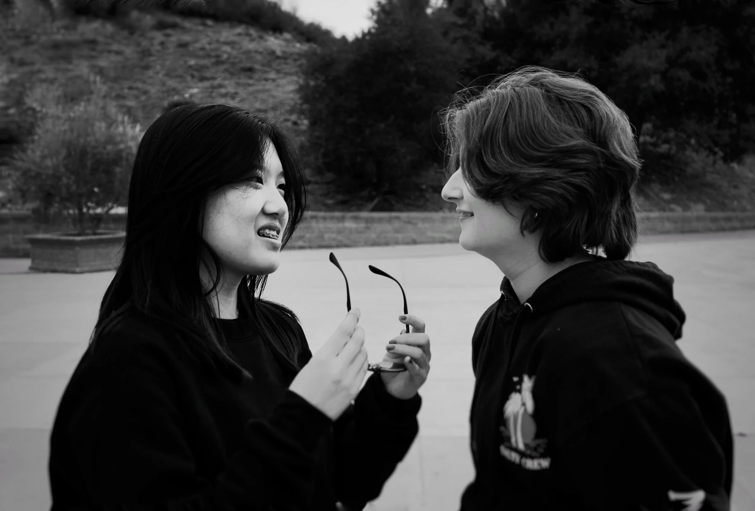 Two young women standing outdoors, one holding sunglasses and smiling, the other looking at her with a smile, in a park with trees and a hill in the background.