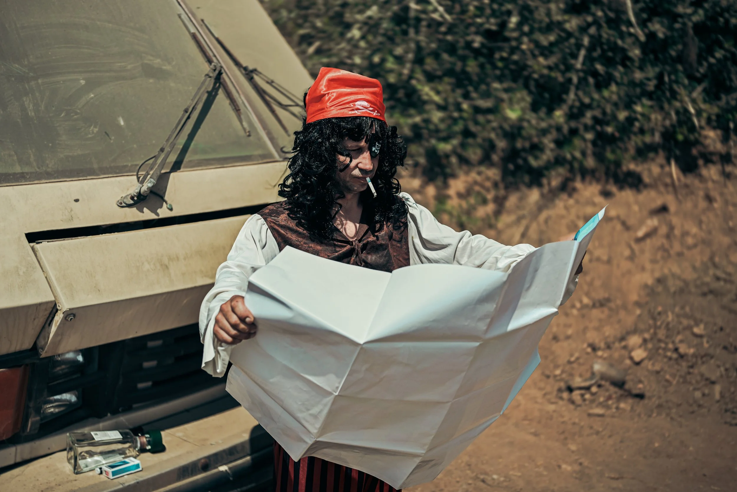 Person dressed as a tourist with a red bandana, sunglasses, smoking, looking at a map outside of an off-road vehicle, in a rugged outdoor setting.