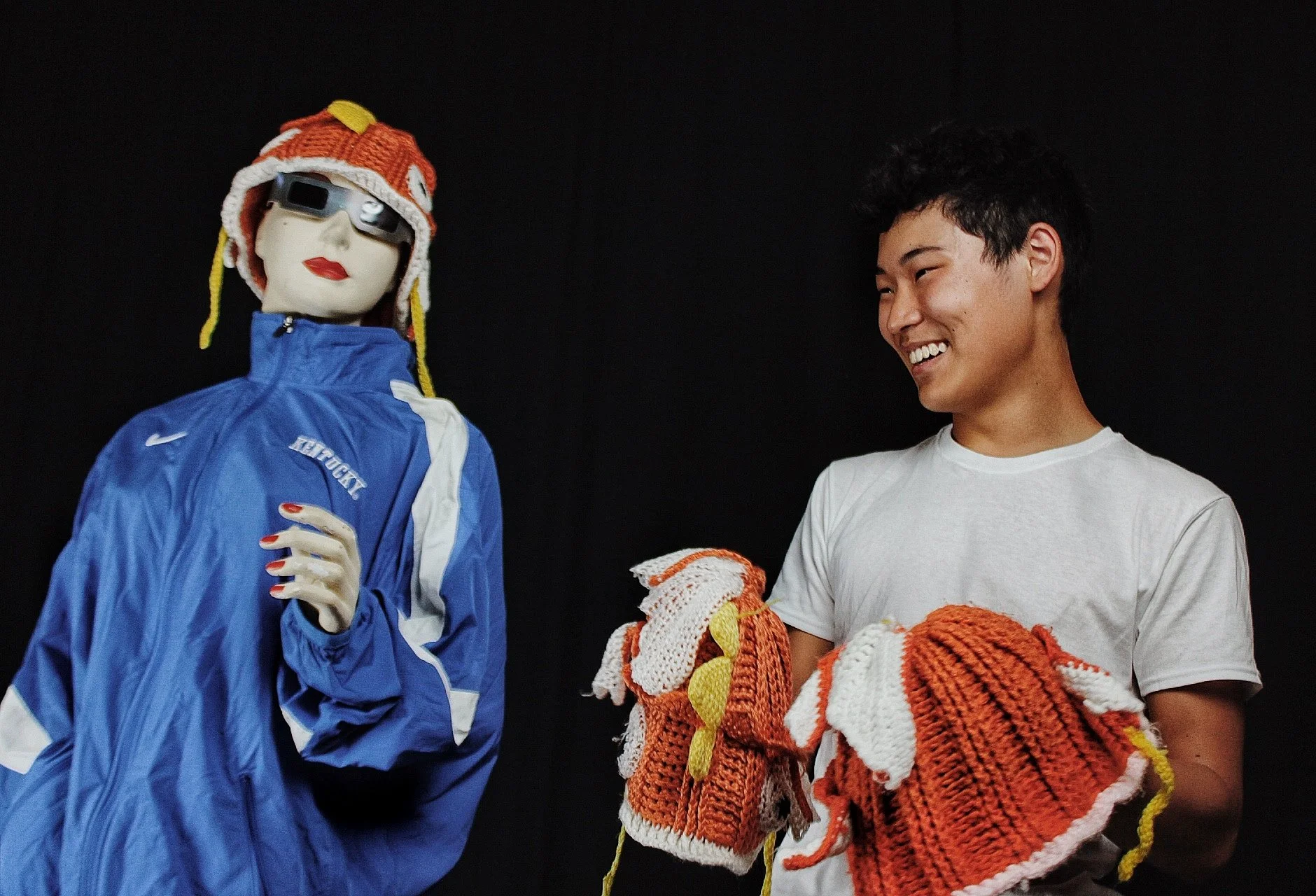 A young man smiling while holding orange, white, and yellow knitted mittens, standing next to a mannequin wearing a blue jacket, a colorful knit hat, and sunglasses, against a black background.