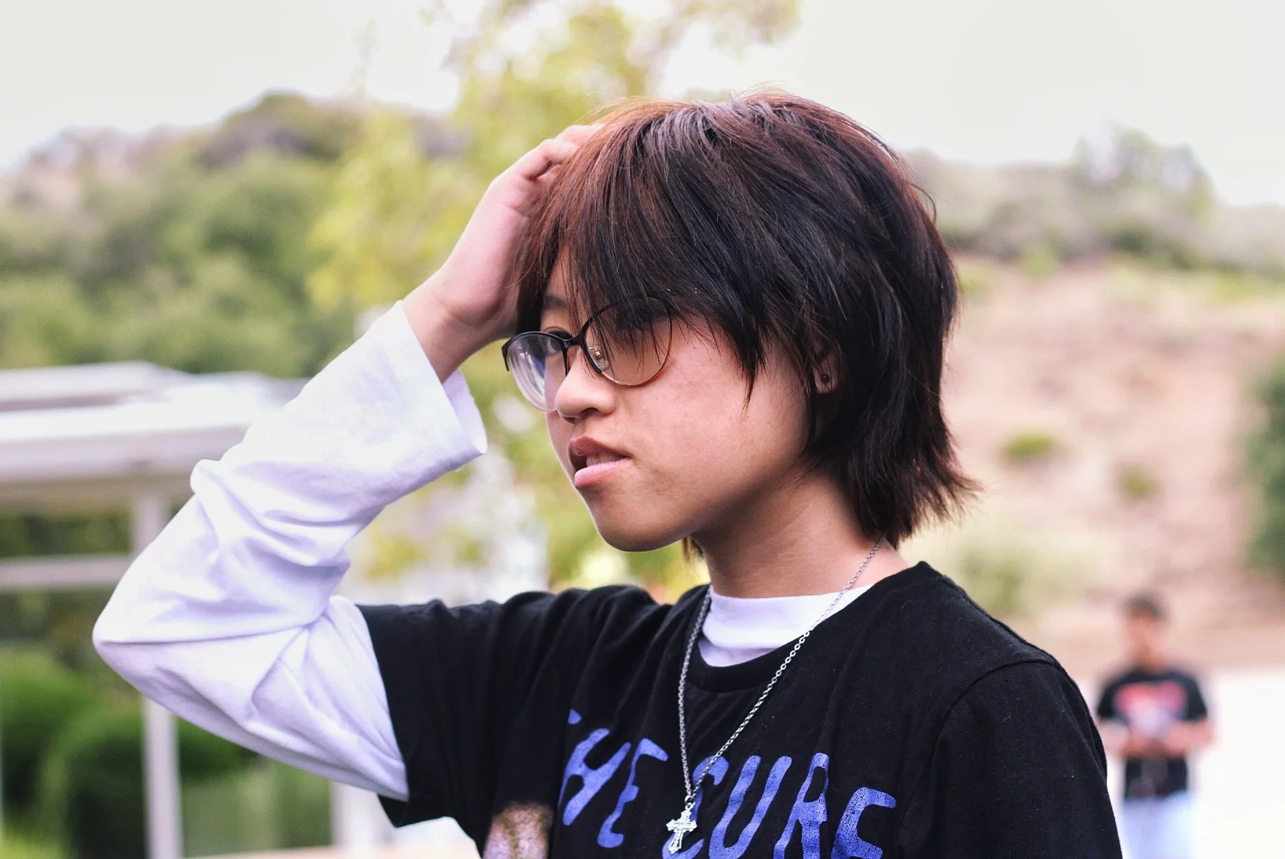 Young person with short, layered hair and glasses, touching their head outdoors.