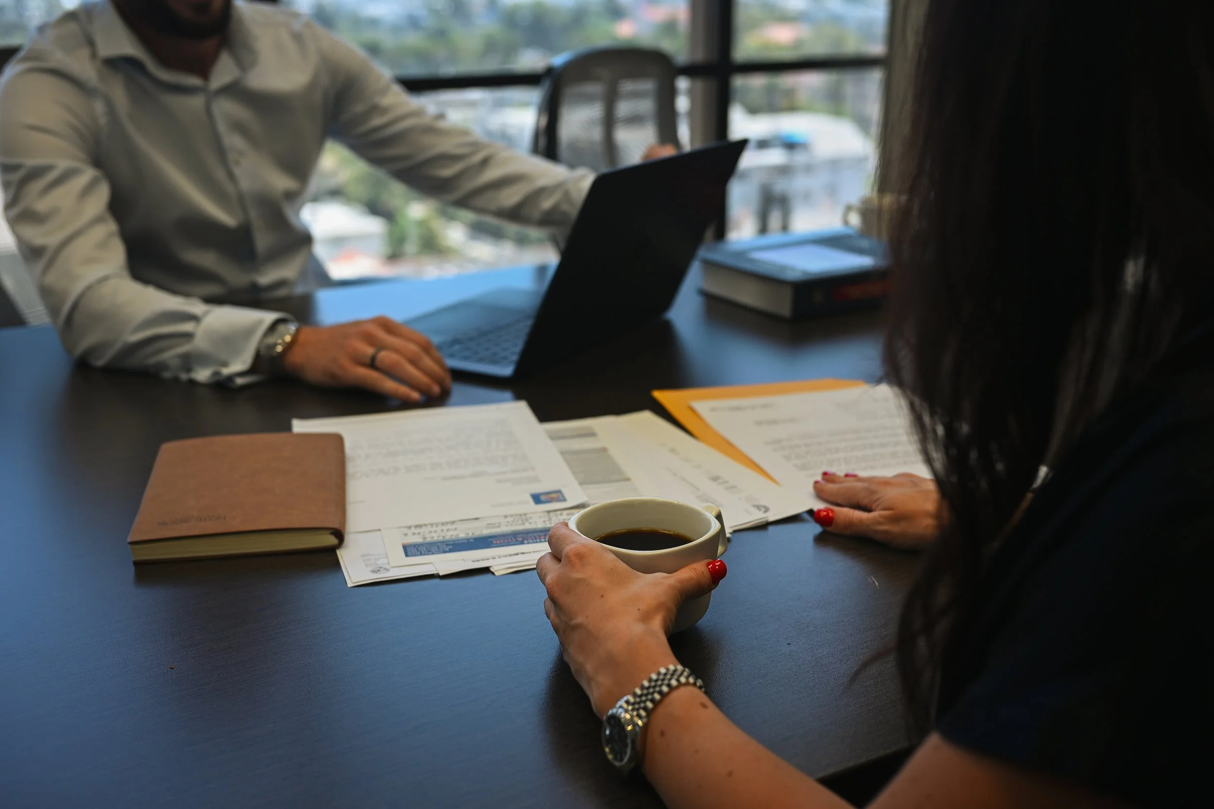 Two people sitting at a table in a meeting room, one woman holding a cup of coffee and the man using a laptop, with documents and books on the table.