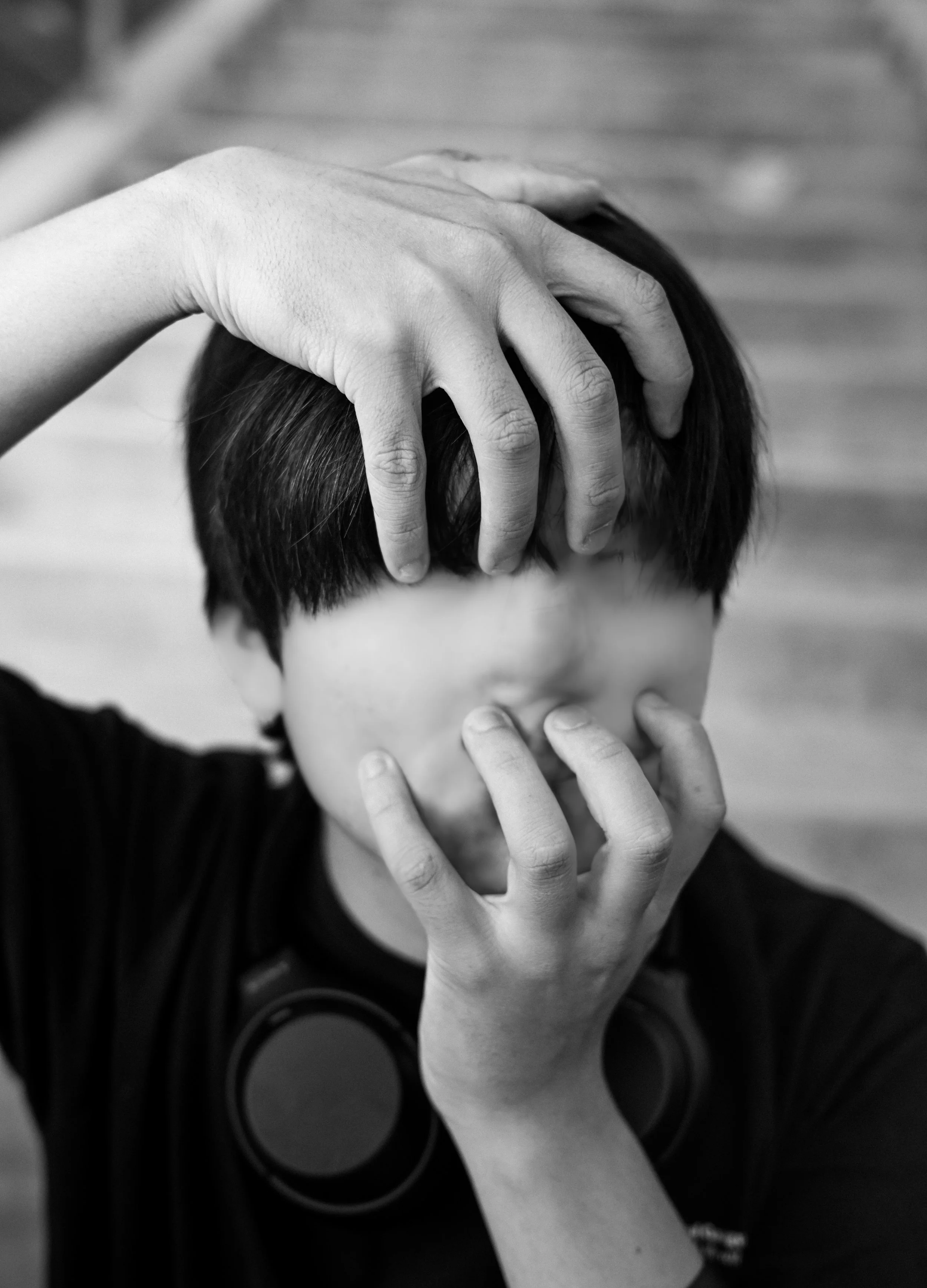 A person with short dark hair covering their face with their hands.