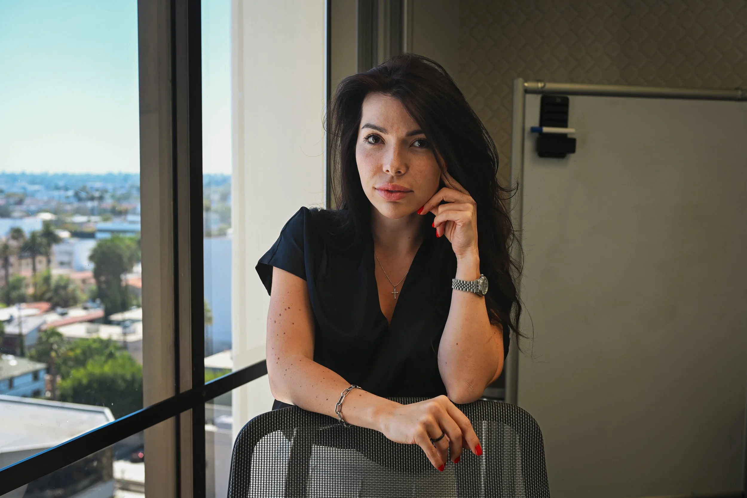 A young woman with dark hair, wearing a black blouse, sitting at a desk with a city view in the background, indoors.