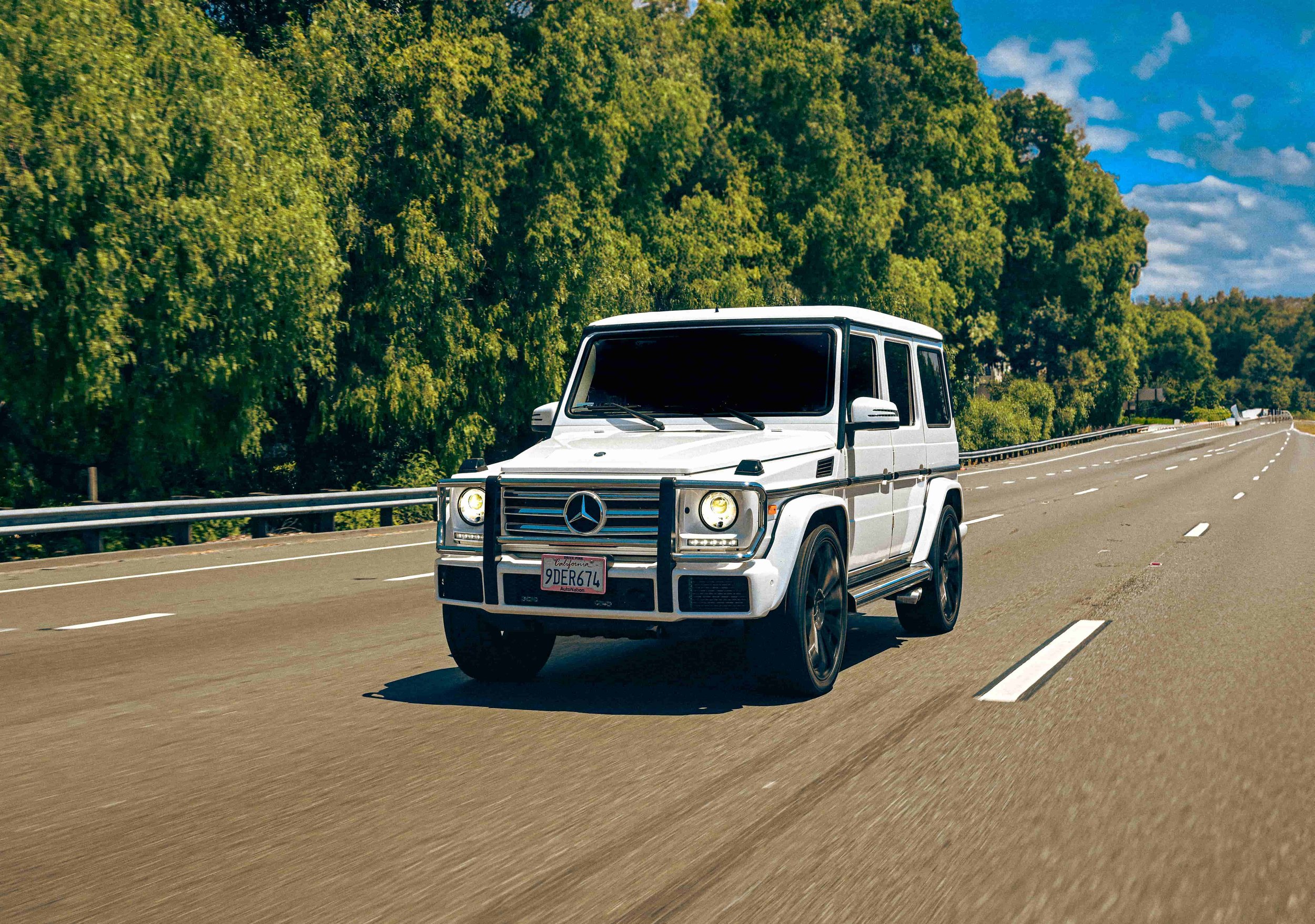 A white Mercedes-Benz G-Class SUV driving on a highway with green trees and a partly cloudy sky in the background.