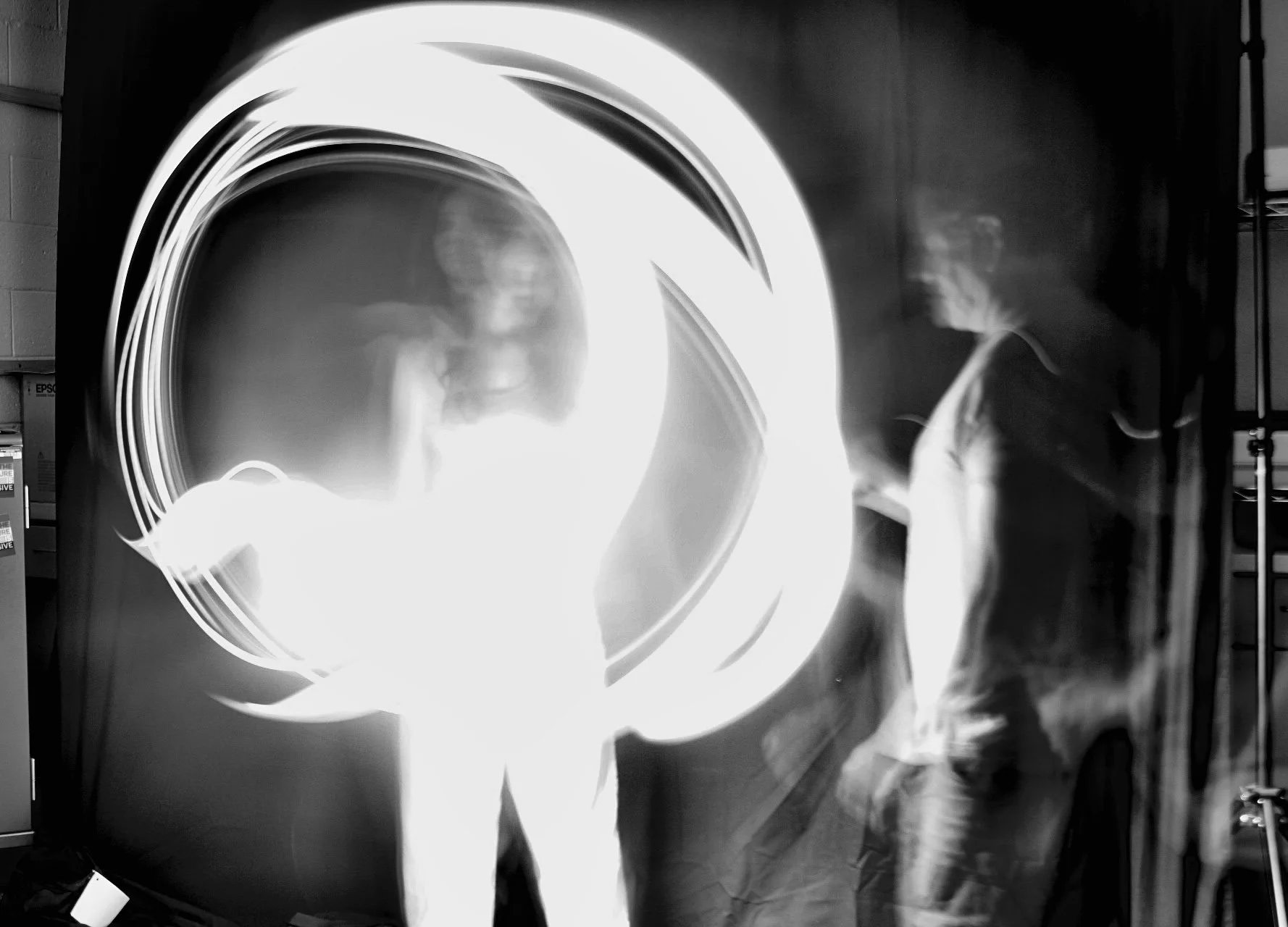 Two individuals standing in front of a bright, circular light display, with reflections on a shiny surface in a dark setting.