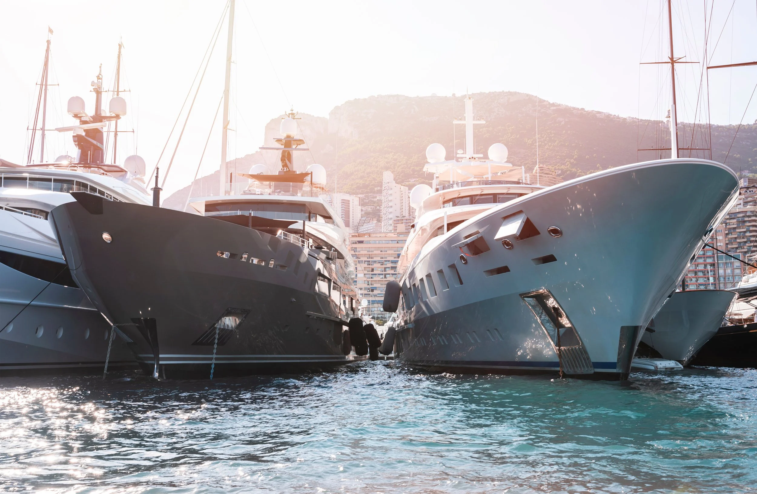 Multiple large yachts docked at a marina with mountains and city buildings in the background.