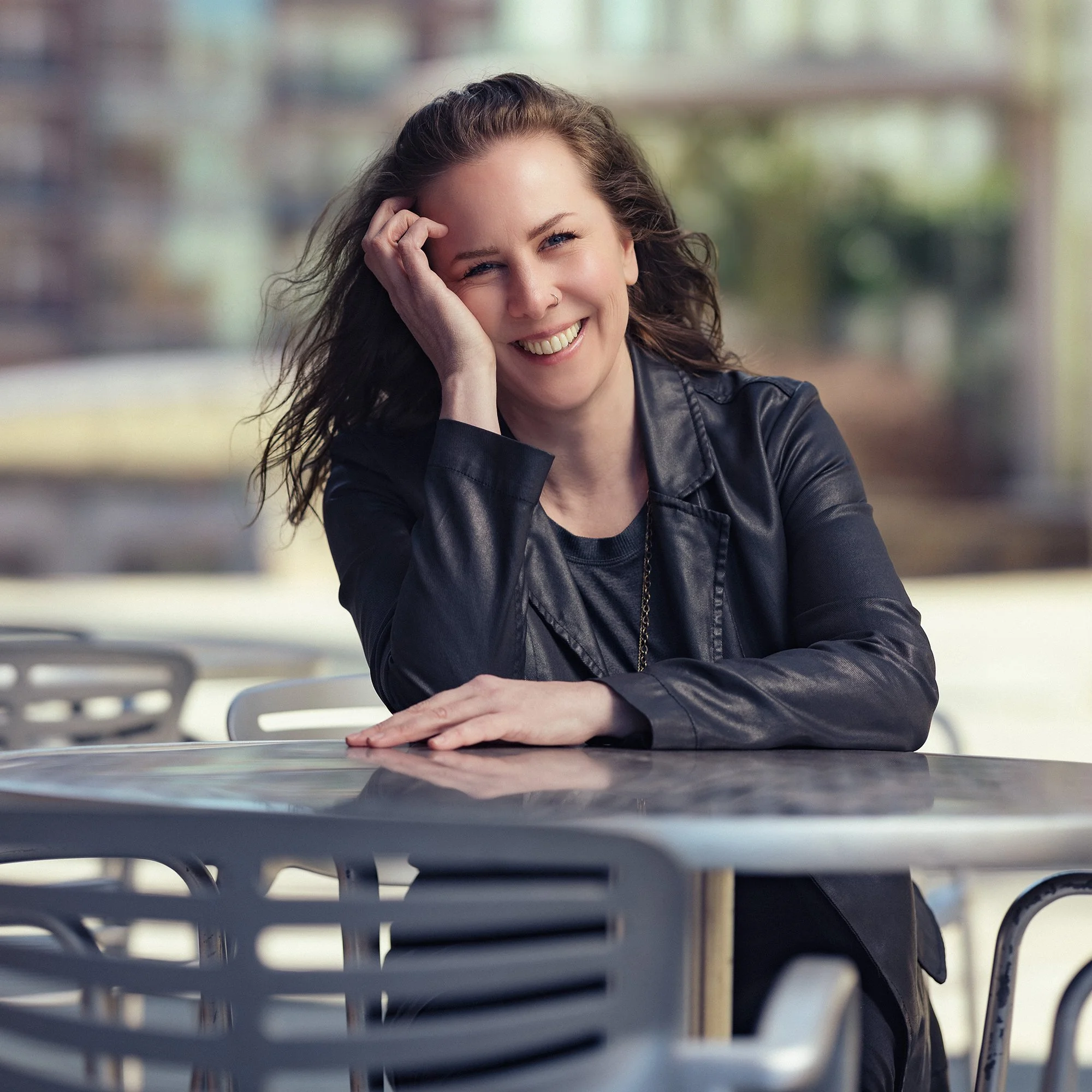 Woman with curly hair smiling, sitting at an outdoor table, wearing a black leather jacket.