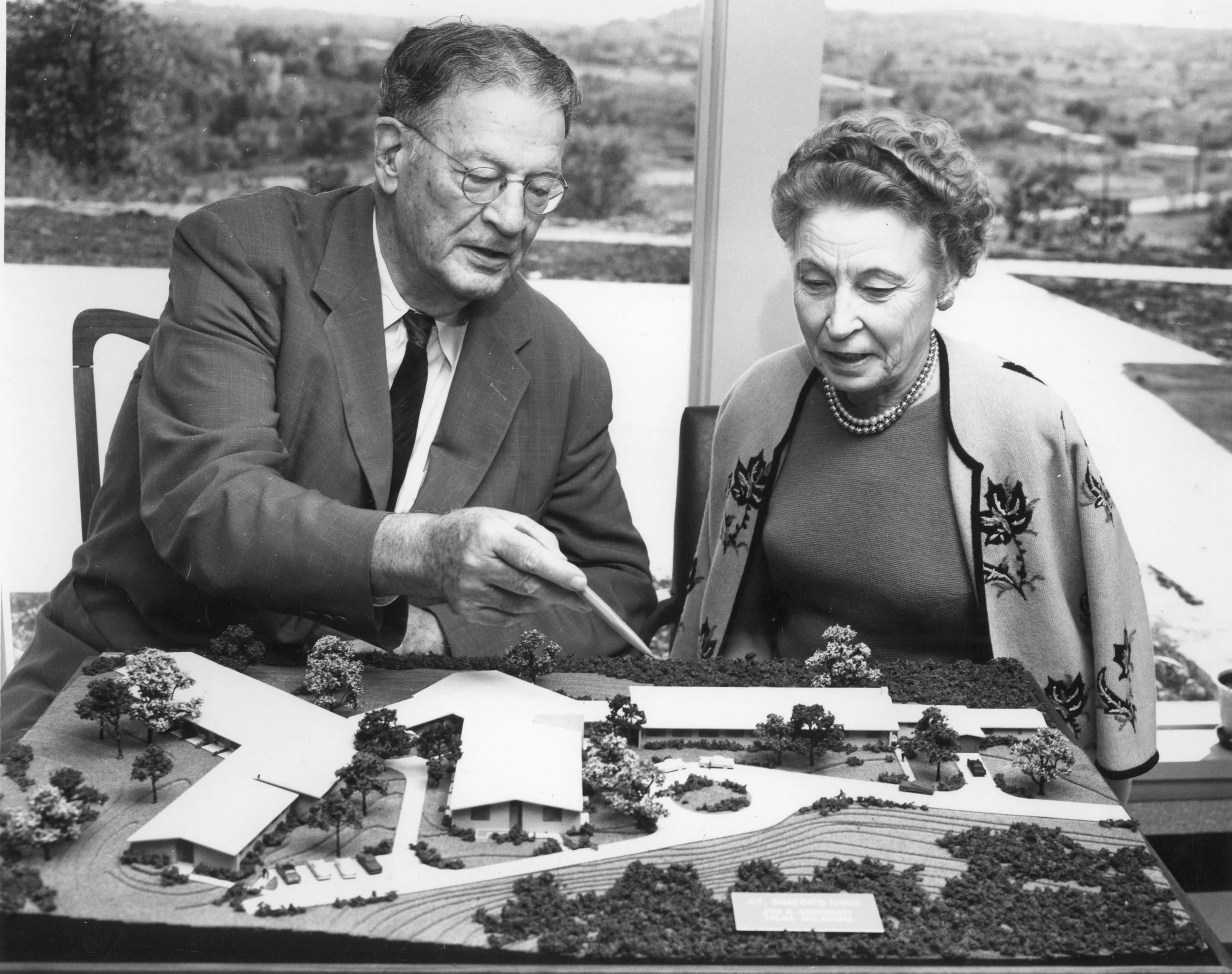 Dr. Eckel and his wife look at an architectural model of the original Saint Simeon's building complex with trees, with a window showing a landscape in the background.