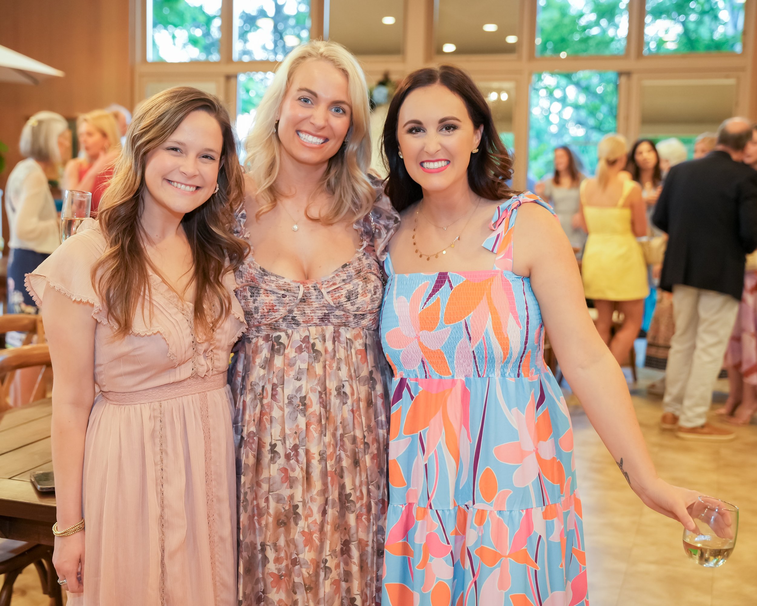 Three women smiling and posing together at an indoor social event, with others in the background.