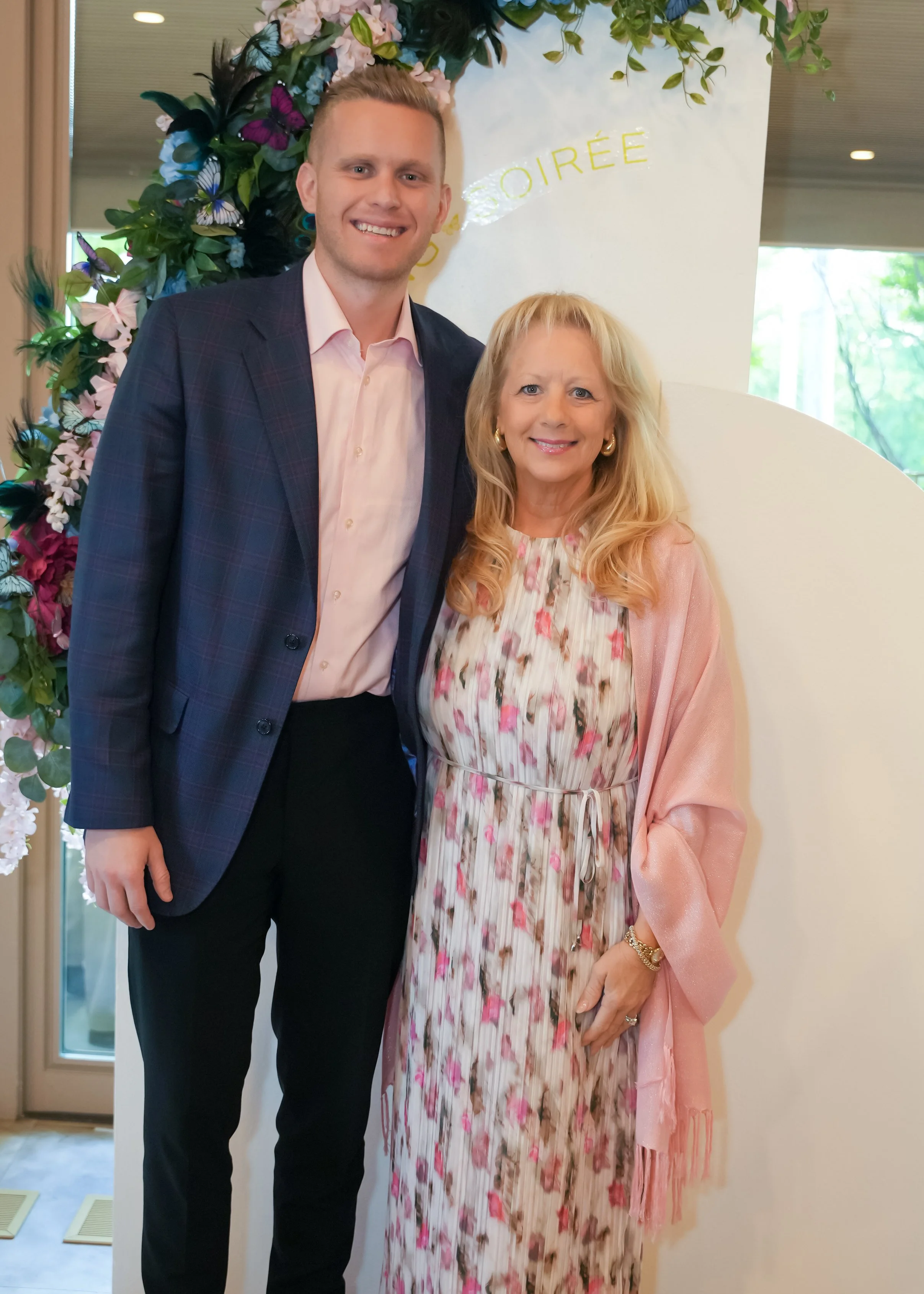 A young man and an older woman standing together at a floral event, smiling at the camera. The young man is wearing a blue plaid blazer and a light pink dress shirt, while the woman is dressed in a floral patterned dress with a pink shawl.