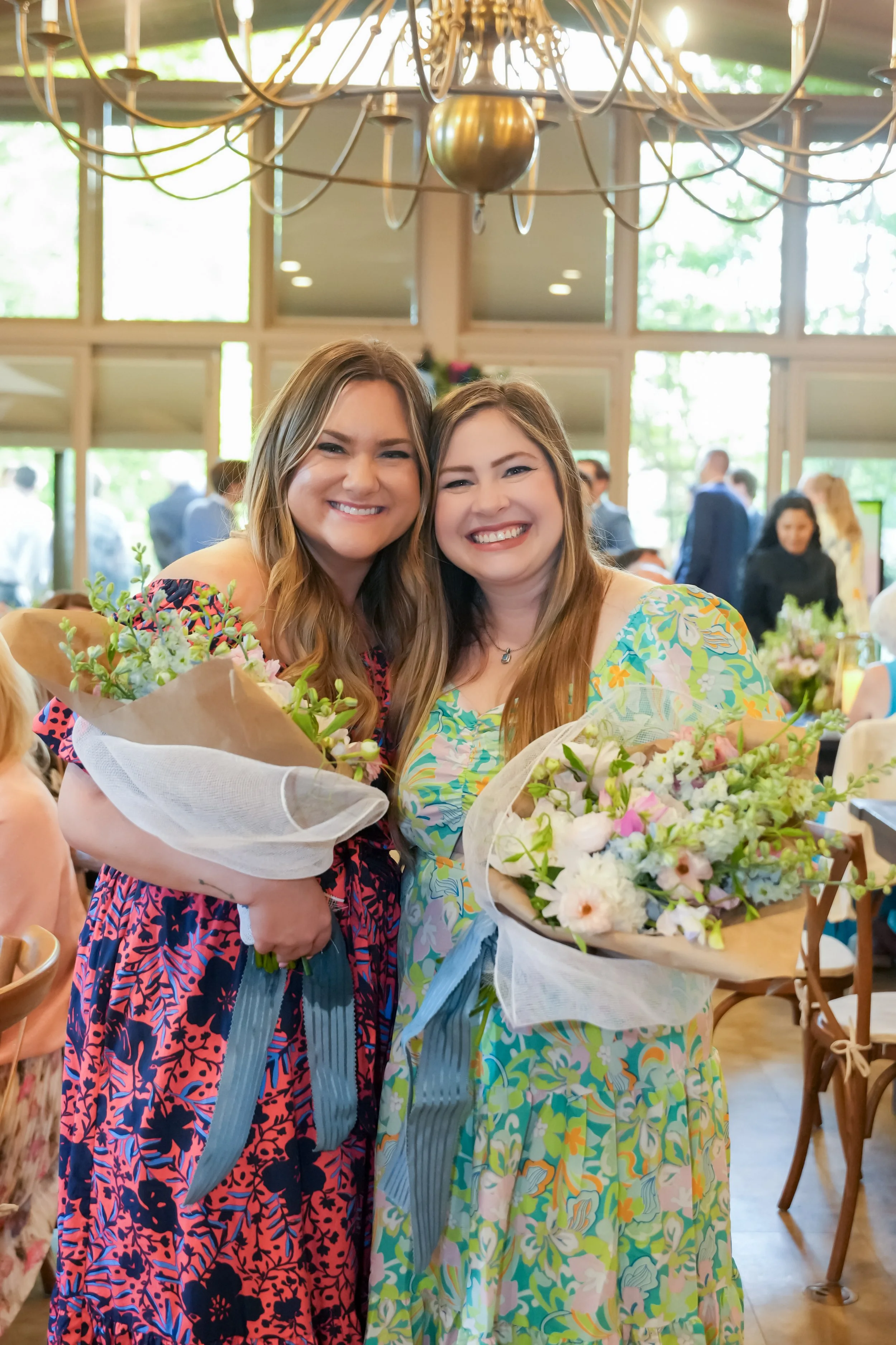 Two women with long hair smiling and holding bouquets at a celebration or event, standing in a bright indoor space with large windows and a chandelier overhead.
