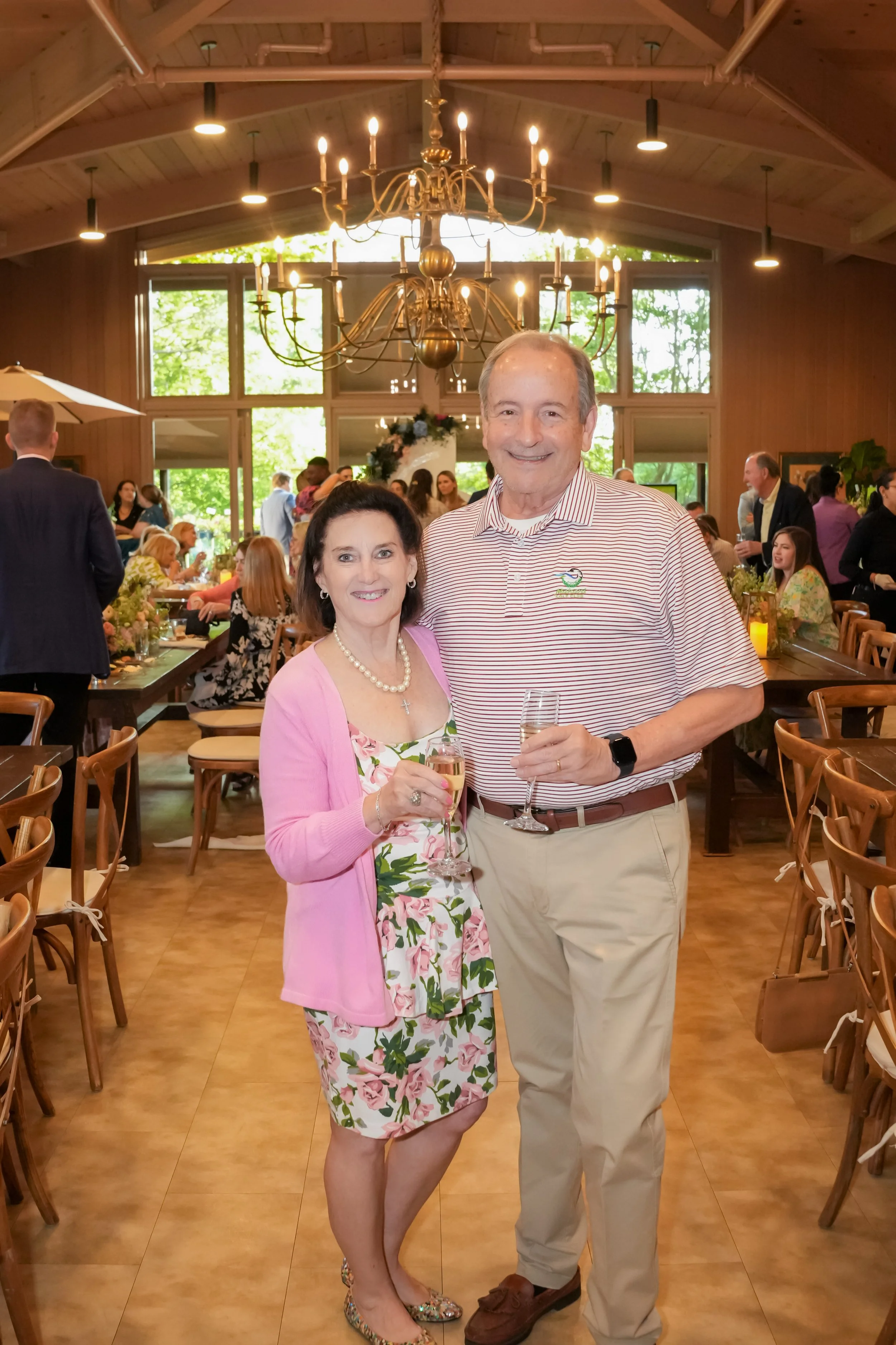 An older woman and man standing together at a social event, holding champagne glasses, in a warmly lit reception hall with many guests.
