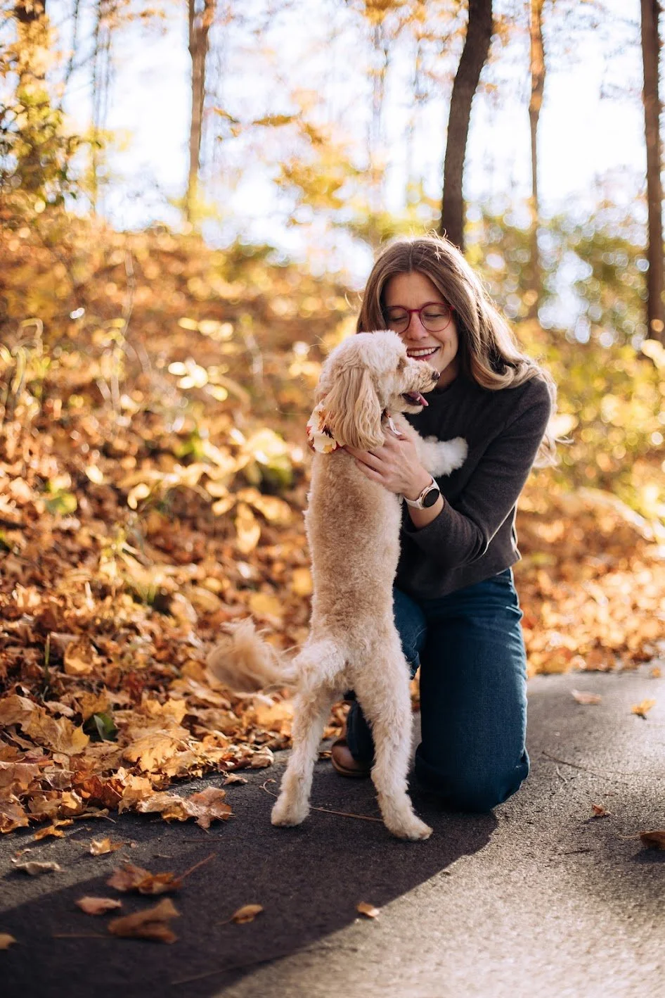 A woman with glasses kneeling on the ground, hugging her dog in a park with autumn leaves.