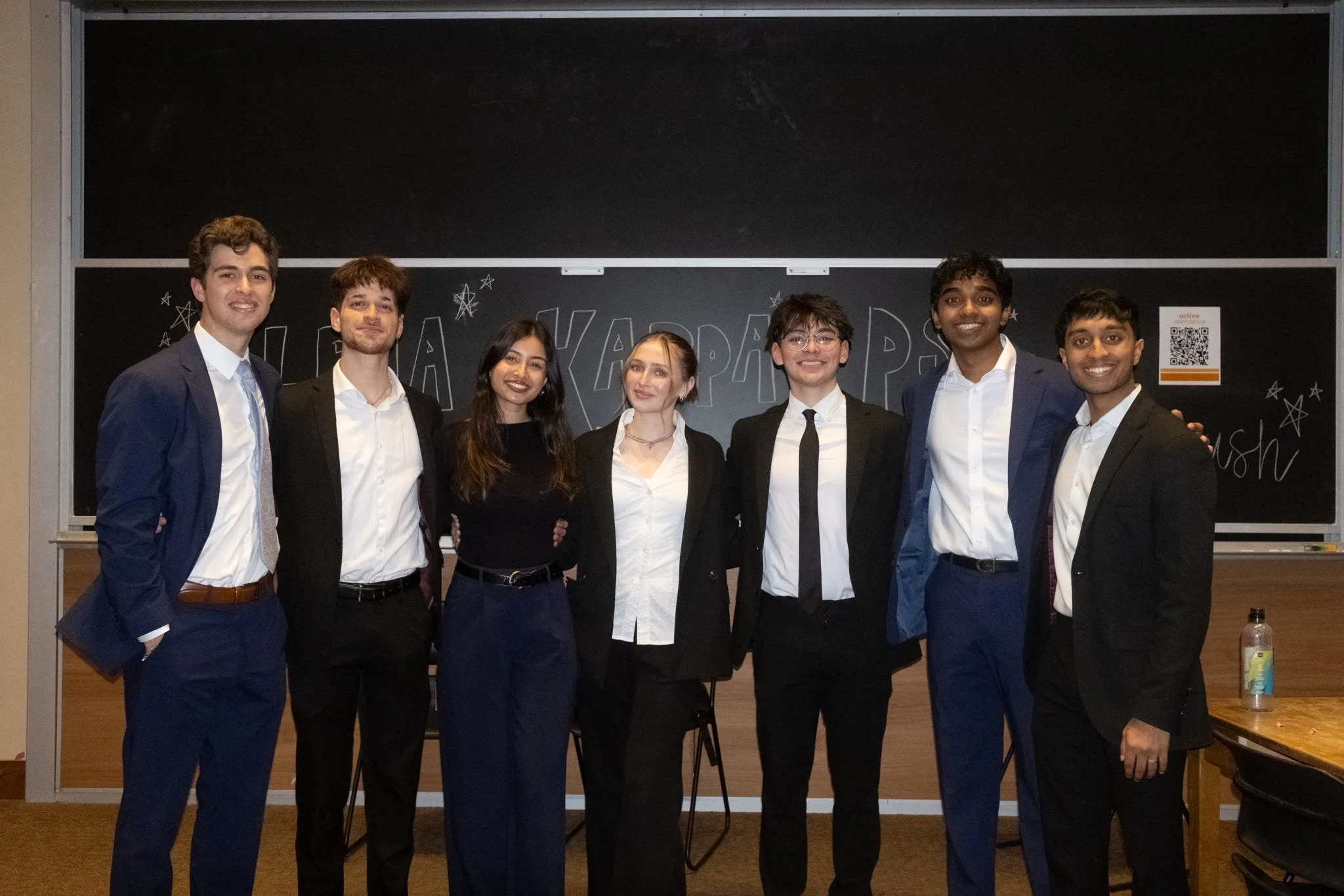 Group of seven young adults dressed in formal attire, standing in front of a blackboard with handwritten text and drawings, smiling for the photo.