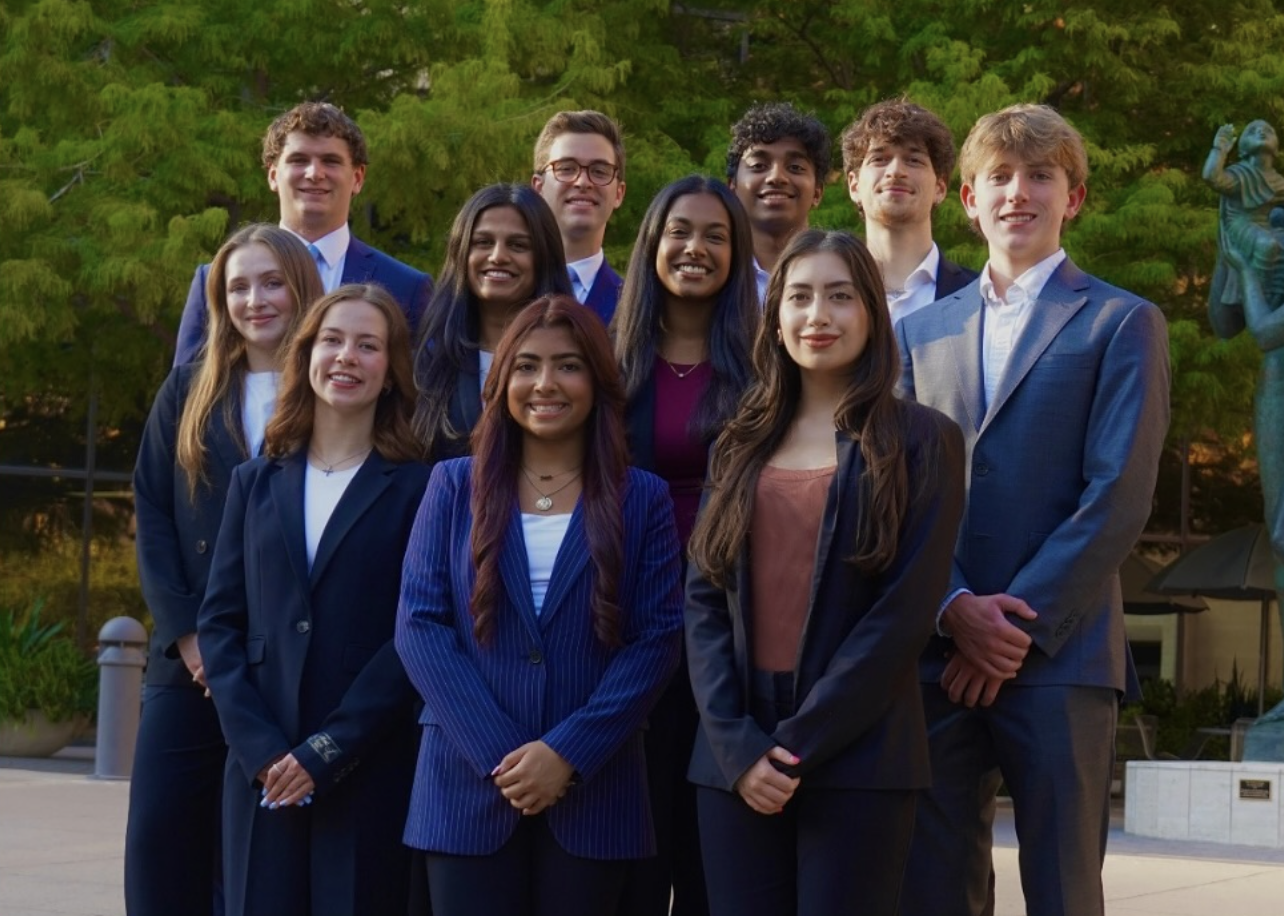 Group of young adults dressed in formal business attire standing outdoors in front of trees and a statue.