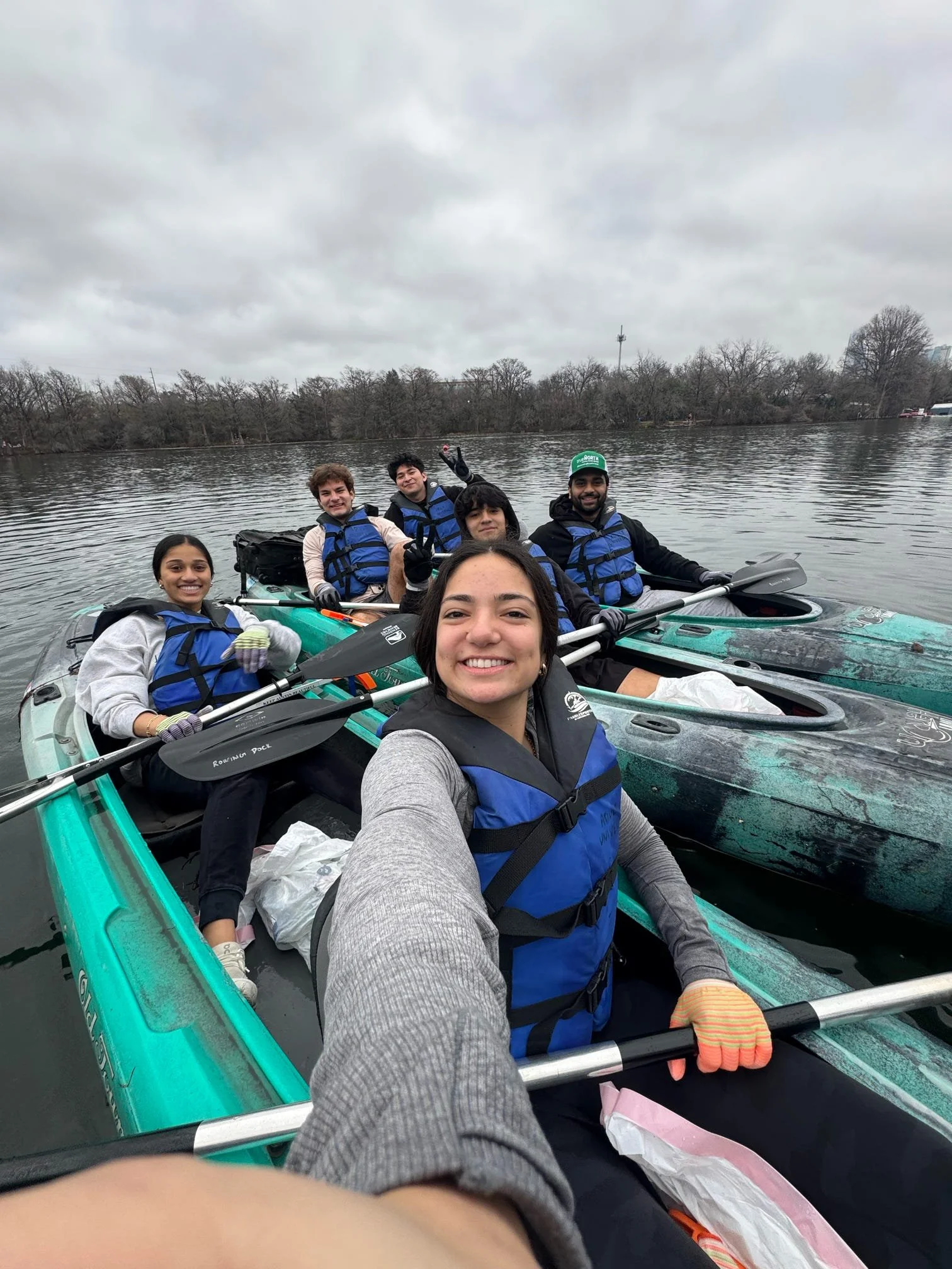 Seven people wearing life jackets on kayaks on a calm river, with a cloudy sky and trees in the background.