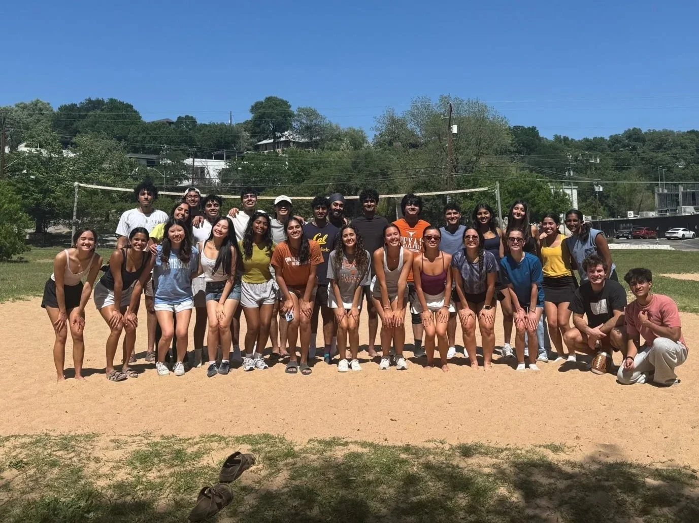 Group of young people standing on a sand volleyball court outdoors, posing for a photo on a sunny day with trees and buildings in the background.