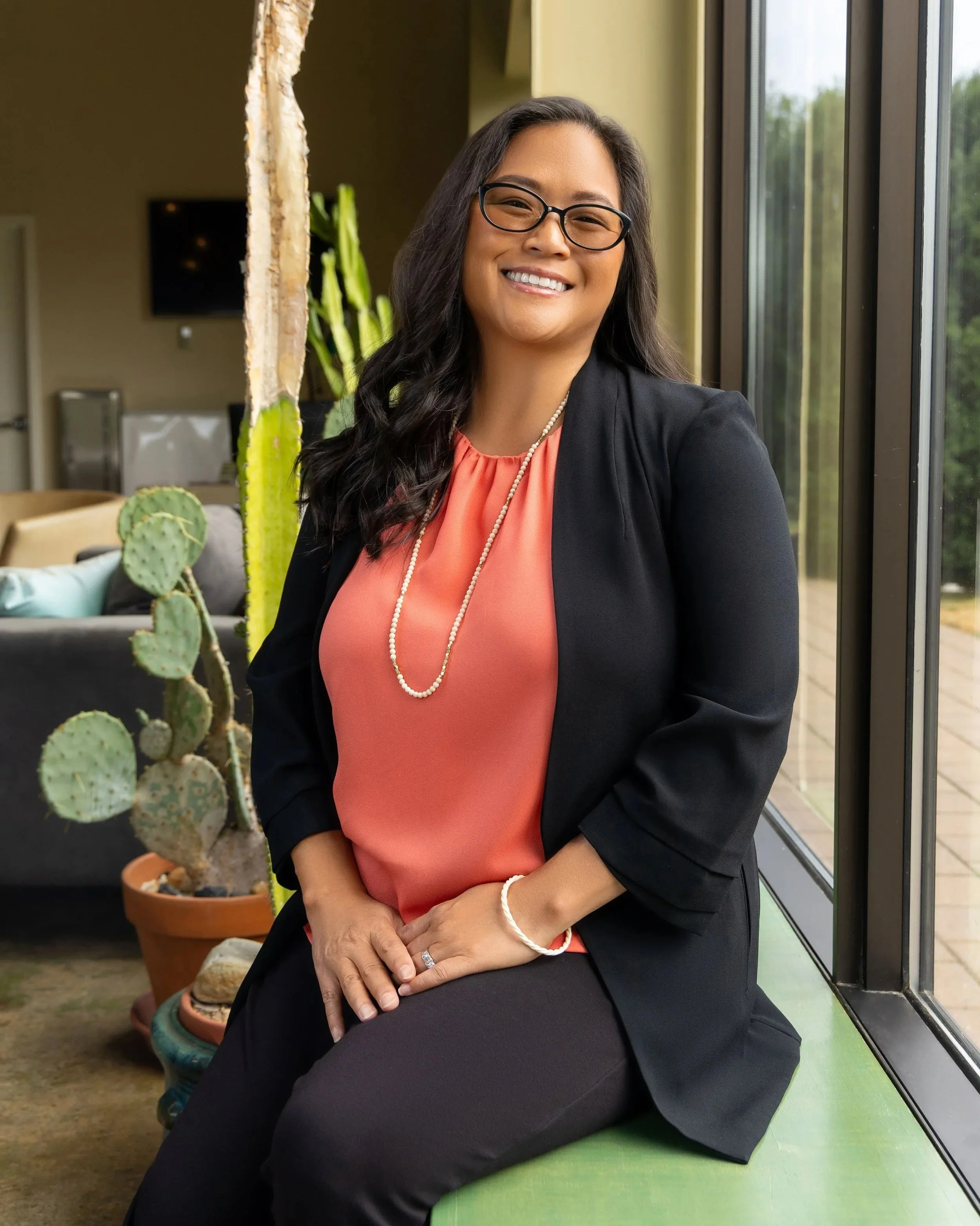 A woman with long dark hair and glasses sitting by a large window, smiling and dressed professionally in a black blazer, coral blouse, and jewelry, surrounded by indoor plants.
