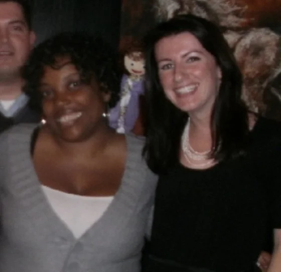 Two women smiling, standing close together, with a woman in the background holding a puppet.