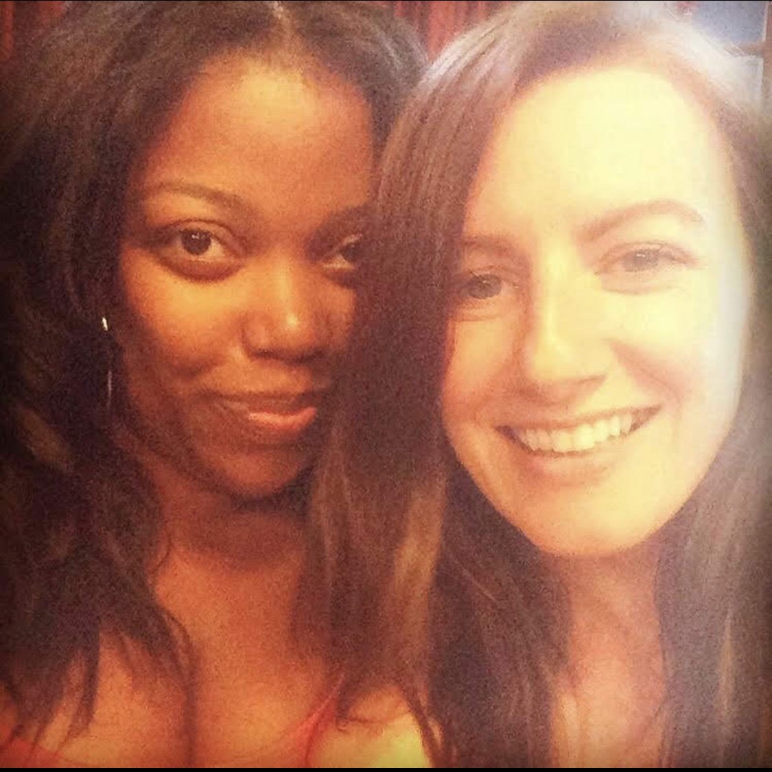 Two women taking a close-up selfie. The woman on the left has dark curly hair and dark skin, the woman on the right has straight brown hair and light skin. Both women are smiling.