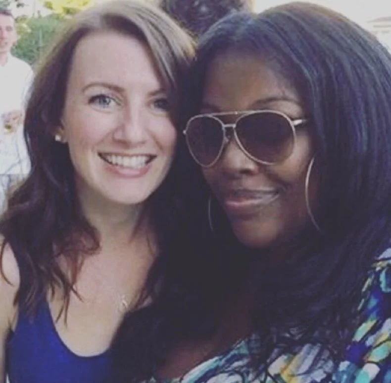 Two women smiling, one with red hair and the other with dark hair wearing large sunglasses, standing close together.