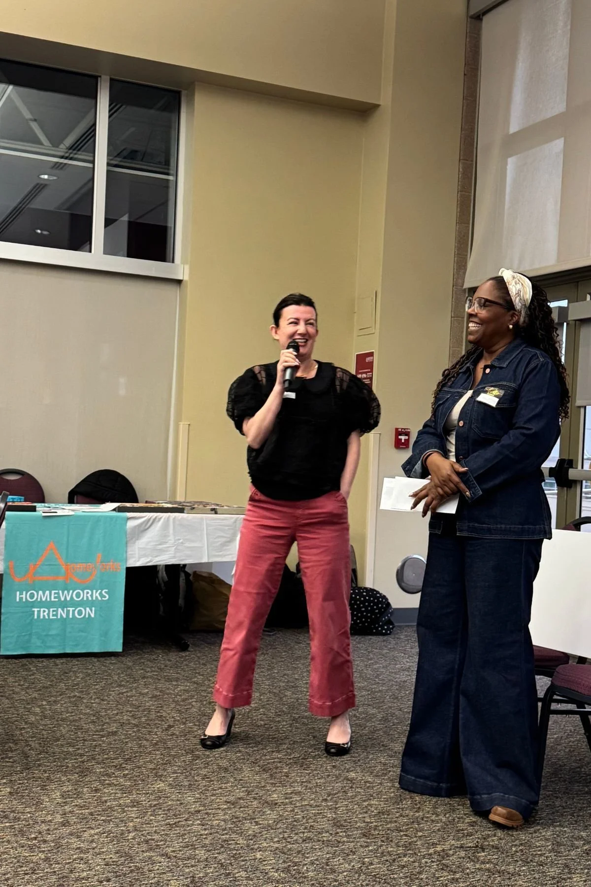 Two women standing and smiling, one holding a microphone, inside a room with a table displaying a banner for Homeworks Trenton.