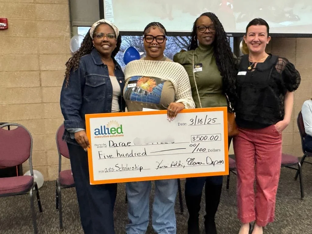 Four women standing together, holding a large check for $500 for a scholarship, inside a room with beige brick walls and chairs, celebrating an achievement.