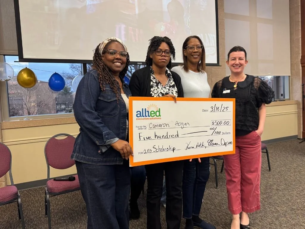 Four women standing indoors, holding a large gift check made out to Cameron Hogan for $500 from Allied executive solutions, with balloons in the background.