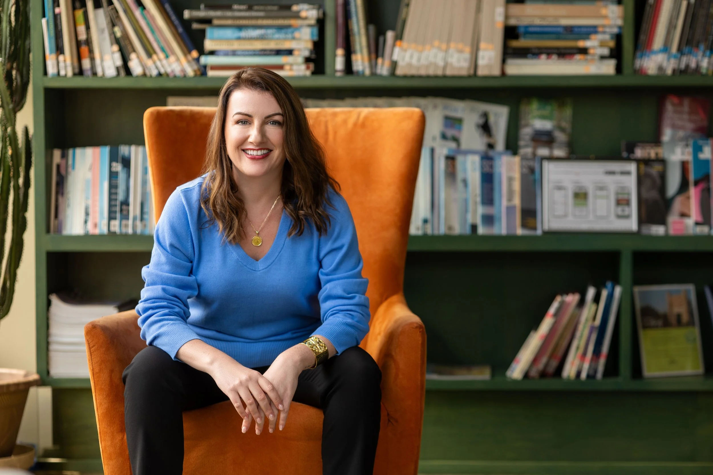 A woman with brown hair wearing a blue sweater, smiling, sitting in an orange armchair in front of a green bookshelf filled with books.