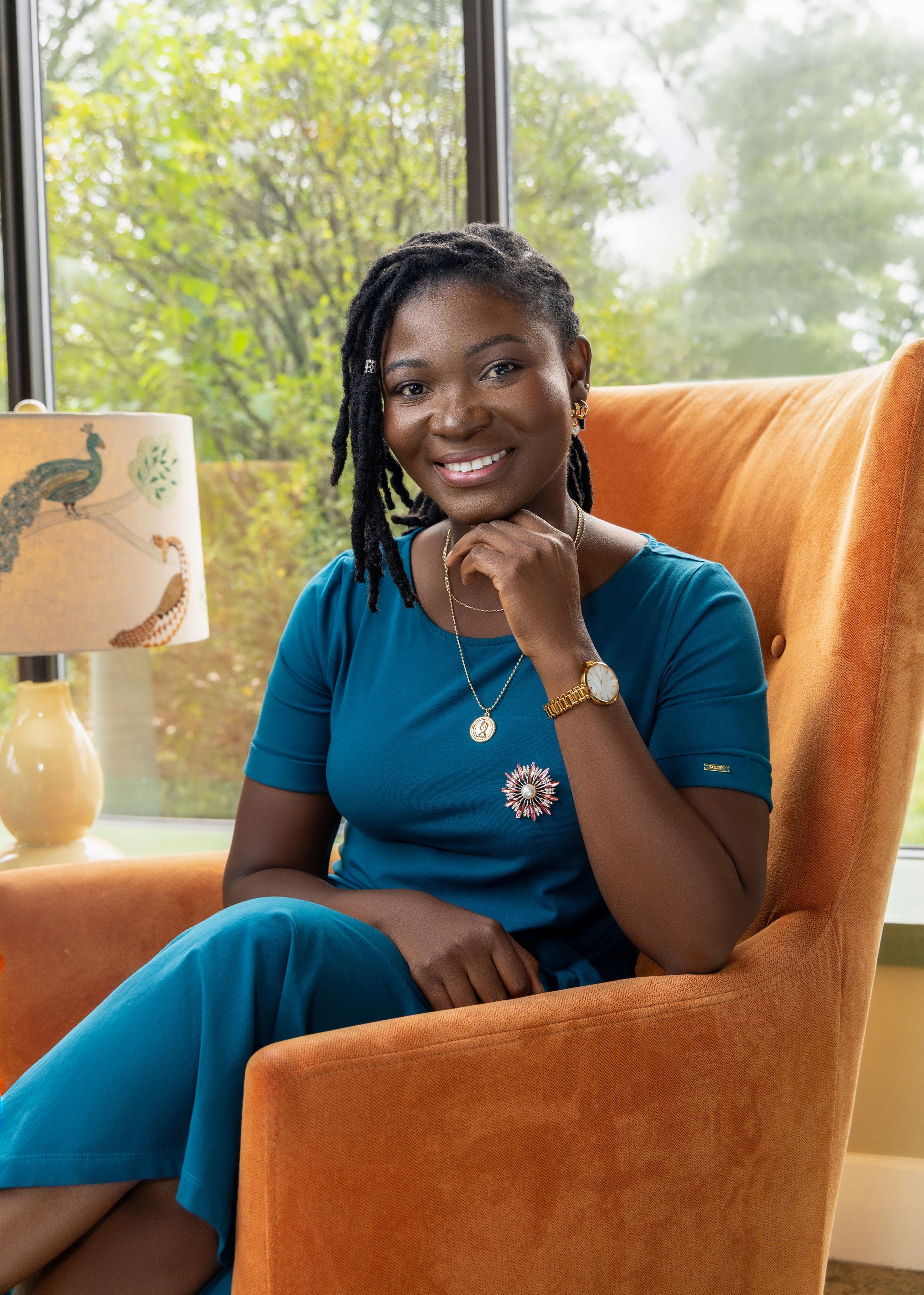 A woman with dark skin and dreadlocks sitting in an orange armchair, smiling at the camera, with a colorful lamp and green trees outside the window in the background.