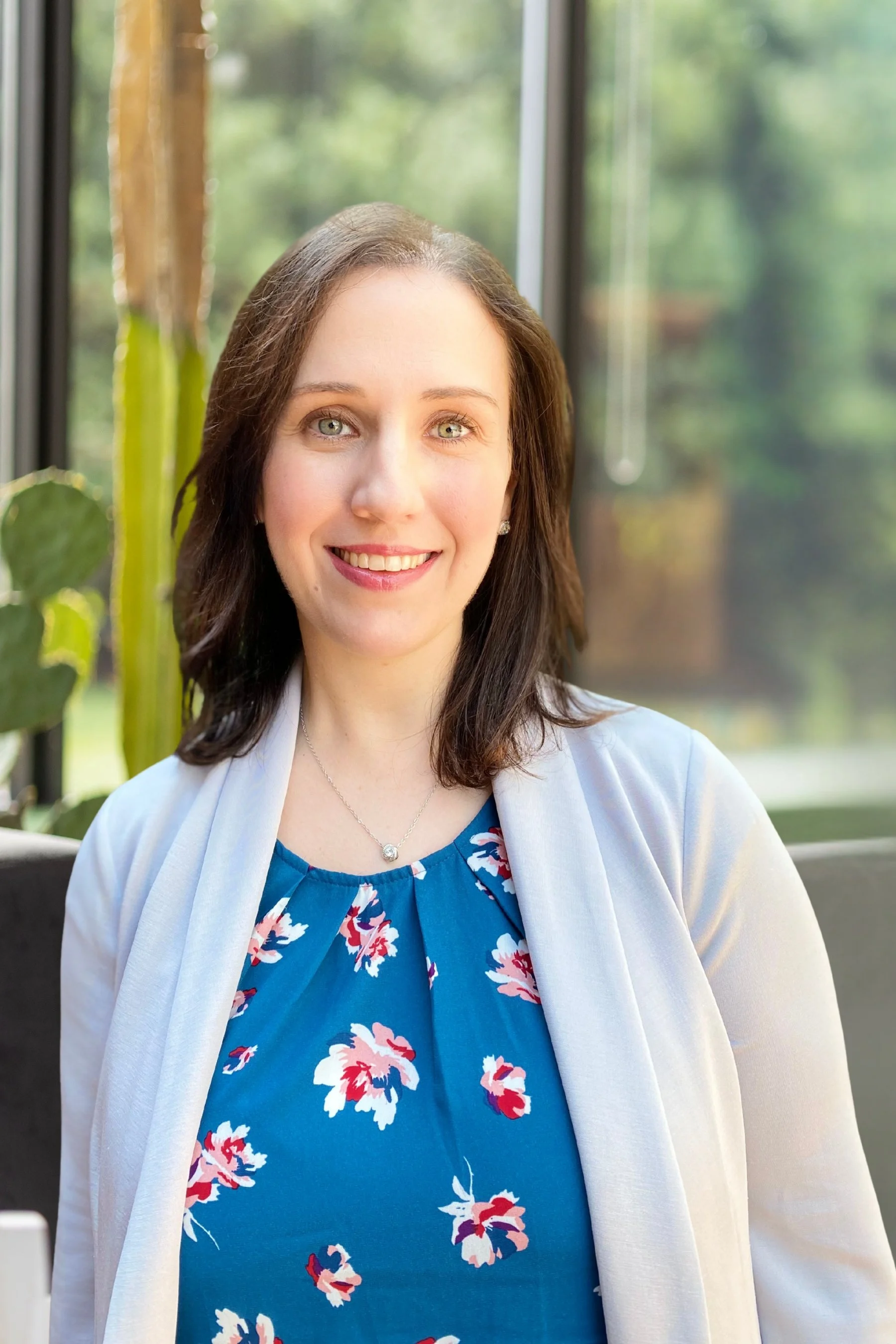 A woman with shoulder-length dark brown hair, wearing a blue dress with a floral pattern and a white cardigan, smiling indoors with green outdoor scenery visible through the window behind her.