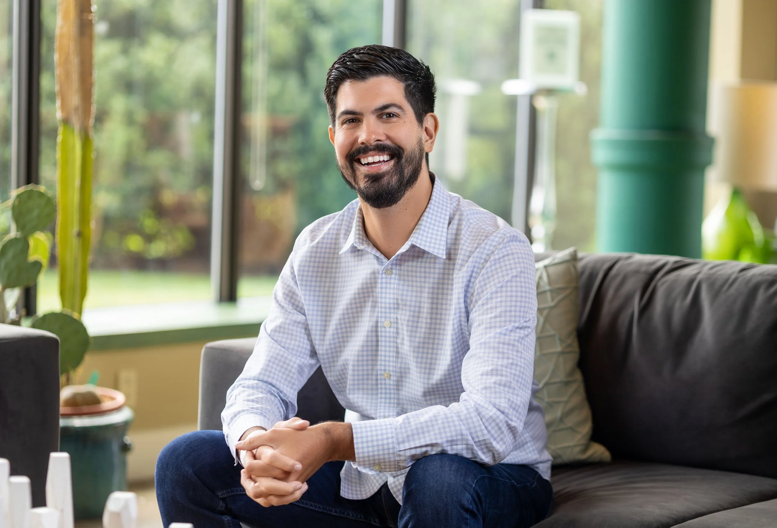 A smiling man with dark hair and a beard, wearing a light blue checkered shirt, sitting on a couch in a bright room with large windows and greenery outside.