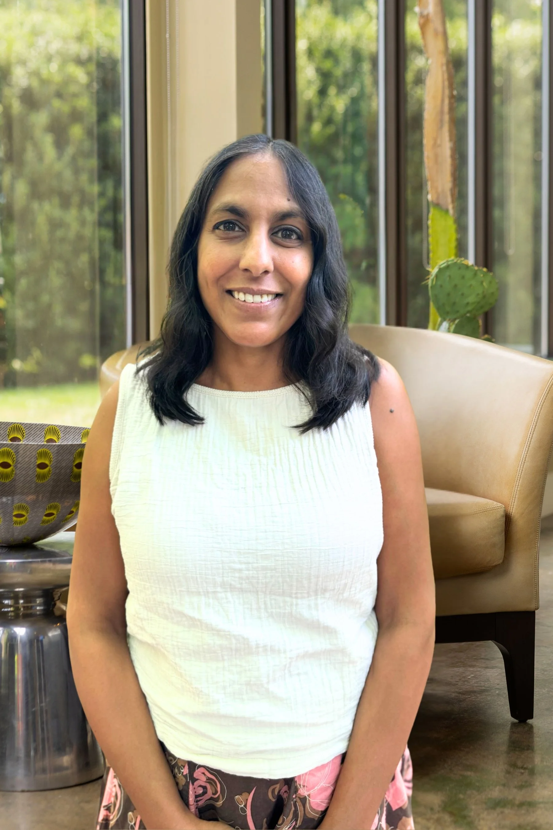 A woman with shoulder-length dark hair, wearing a sleeveless cream-colored top, smiling, seated in a cozy room with large windows showing green trees outside, a cacti plant, and a decorative bowl on a stand.
