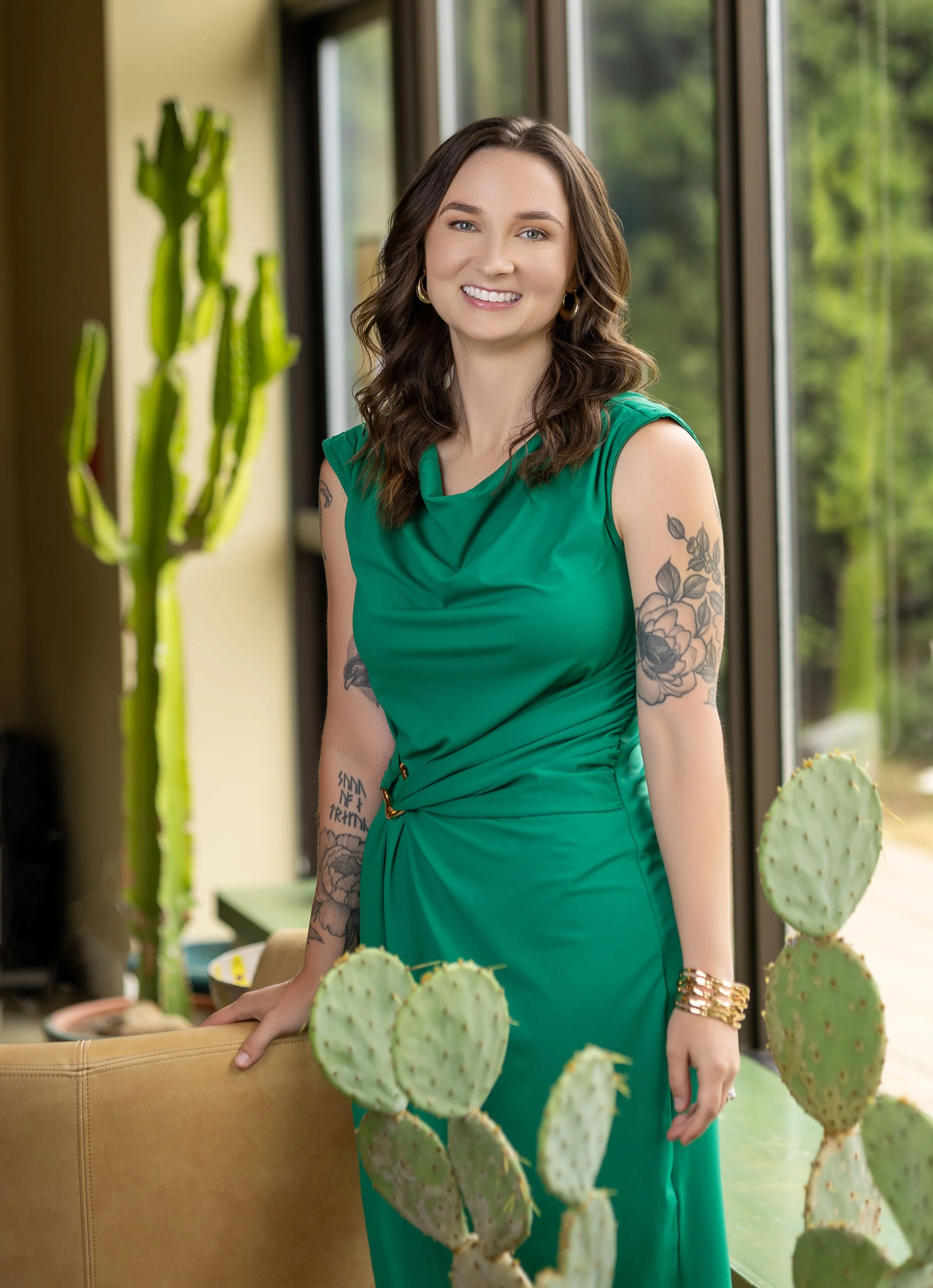 A woman with brown wavy hair smiling, wearing a green sleeveless dress, standing indoors near a beige chair with green cacti in the foreground and large windows in the background.