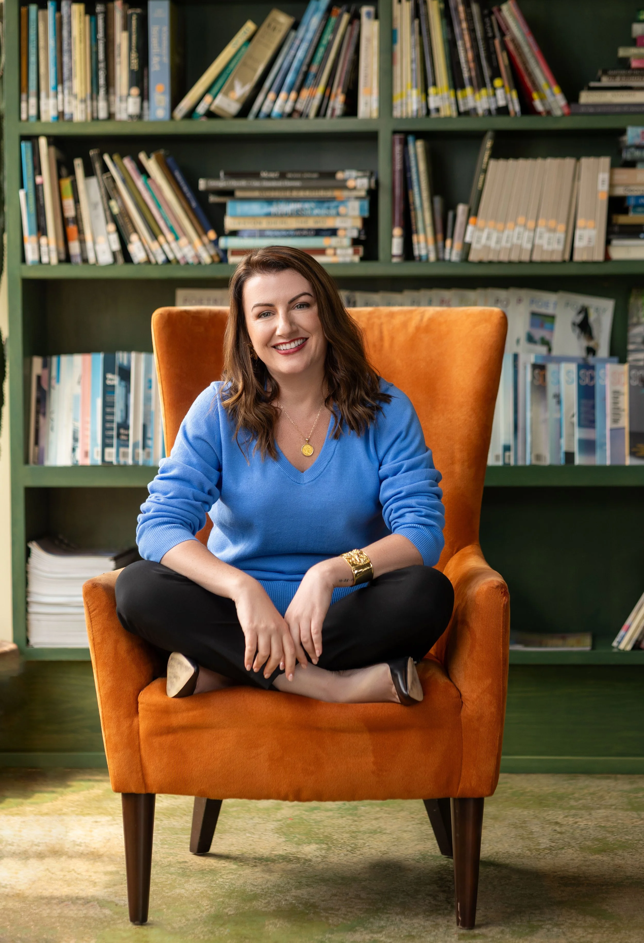 A woman with brown hair, sitting cross-legged in an orange armchair, smiling, in front of a green bookshelf filled with books.