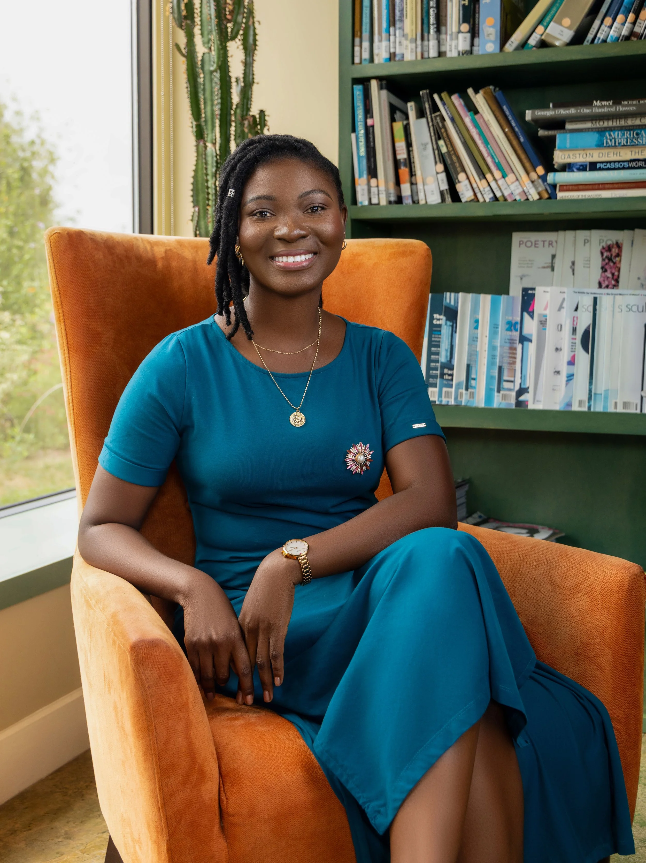 A woman with dark skin and dreadlocks seated in an orange armchair in a library, wearing a blue dress with a brooch on the sleeve, gold jewelry, and a watch, smiling at the camera.