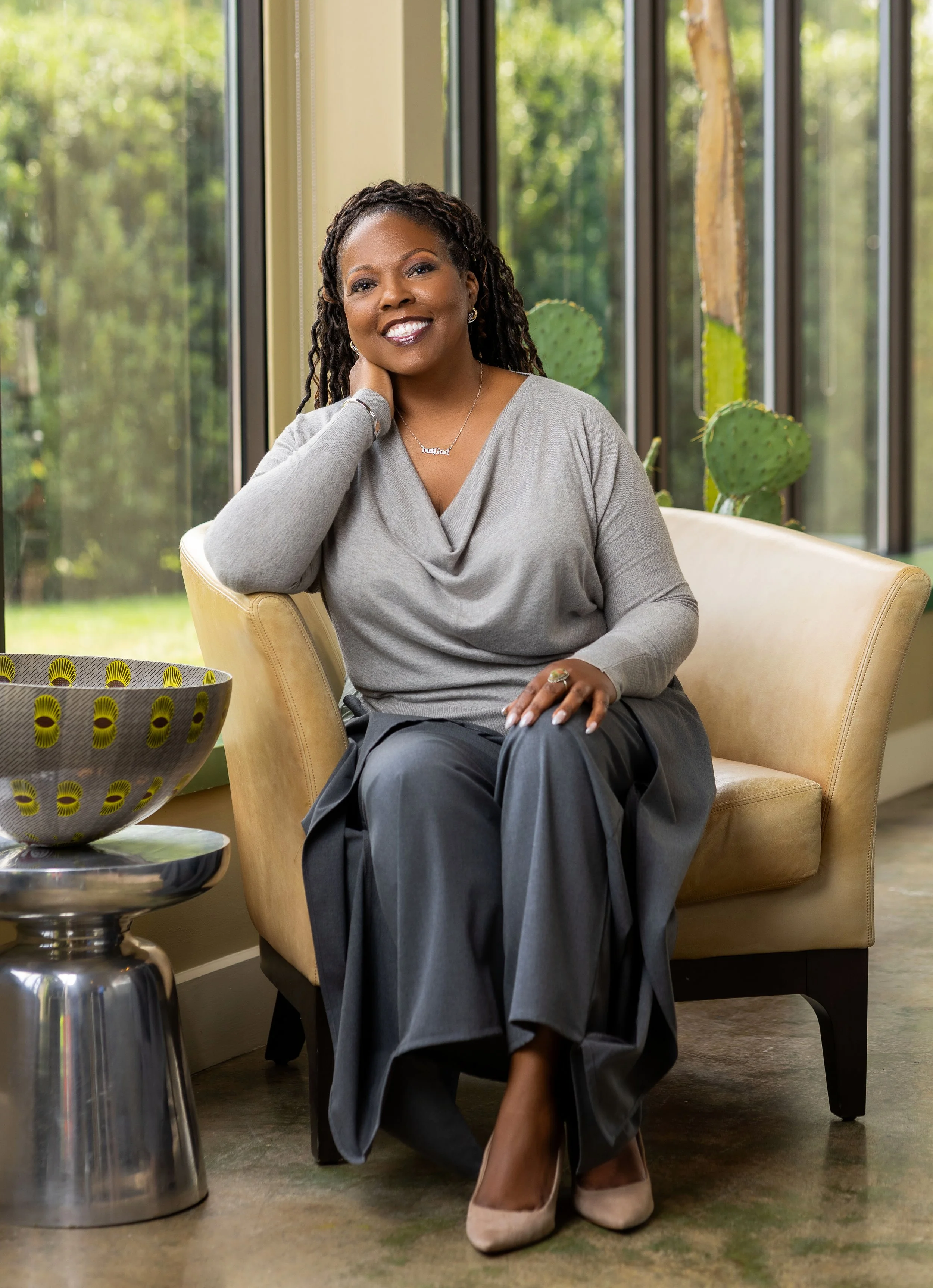 A woman sitting on a beige armchair, smiling, with a cactus plant behind her and a large bowl on a side table, in front of large windows showing a green outdoor landscape.