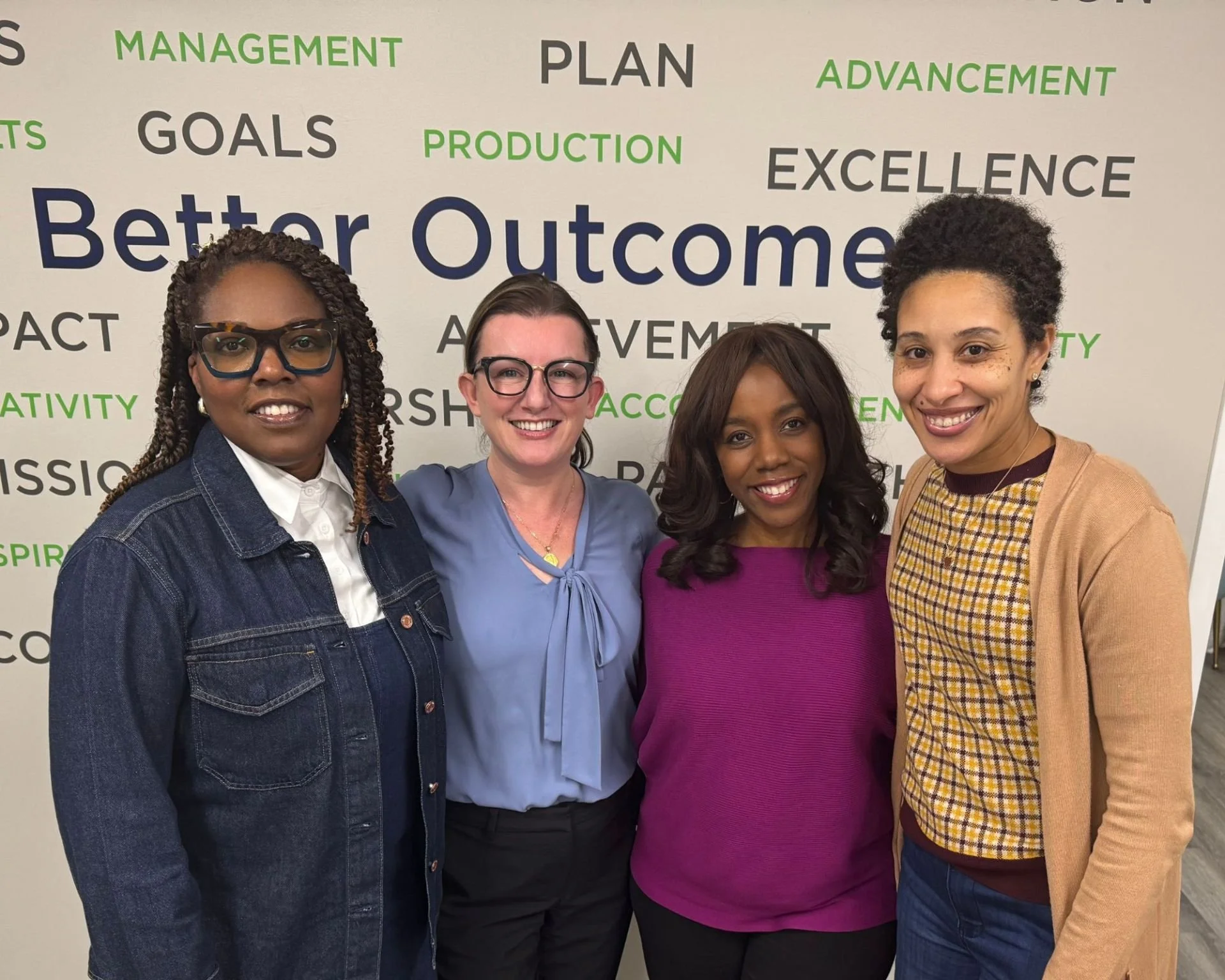 Four women smiling and standing together in front of a wall with words related to management, goals, achievement, and excellence.