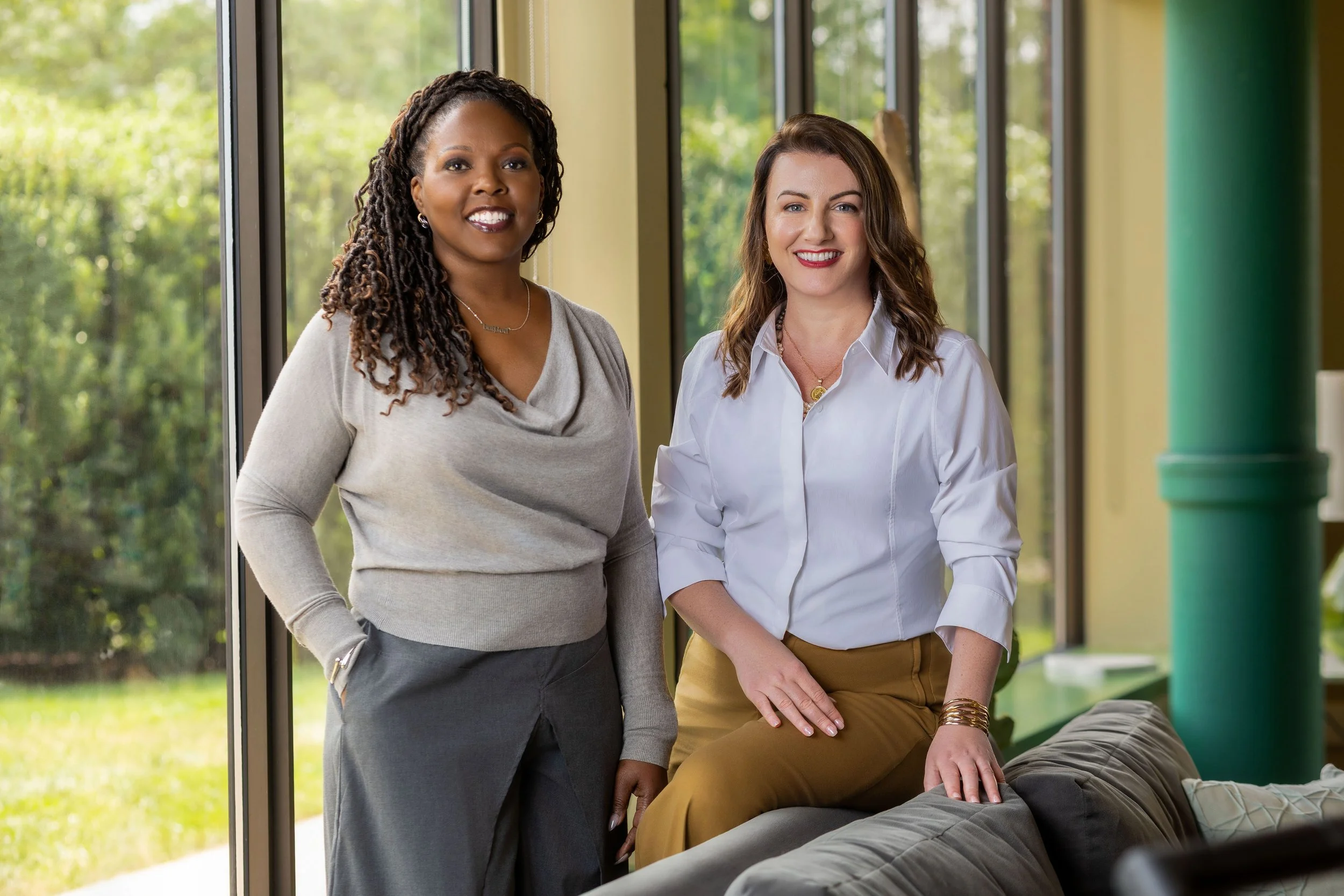 Two women standing inside near large window with greenery outside, smiling, one sitting on the arm of a couch, wearing business casual attire.