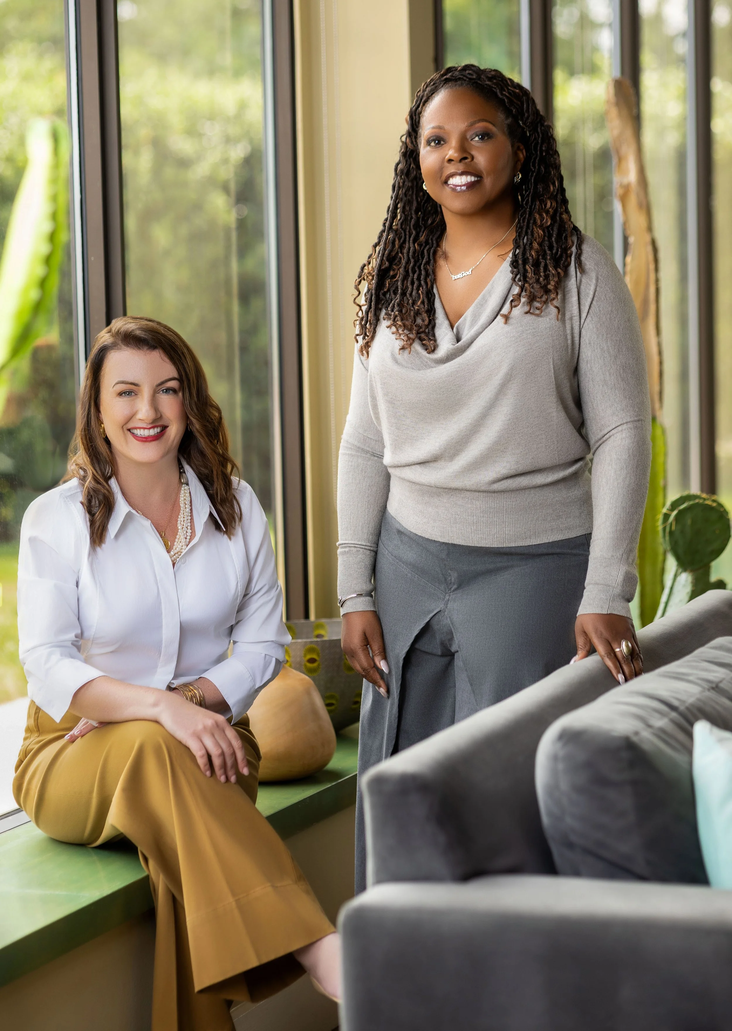 Two women, one sitting on a windowsill and one standing beside her, posing inside a modern room with large windows and green outdoor scenery.